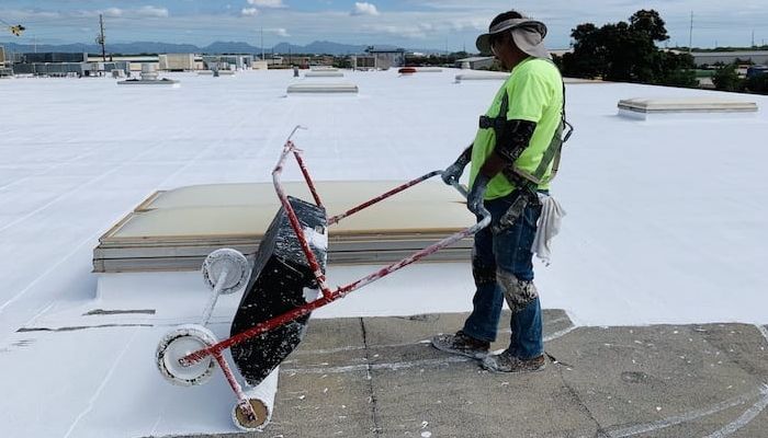 Roofer in green shirt, safety gear using a rolling tool on a white roof. Sunny outdoor setting.