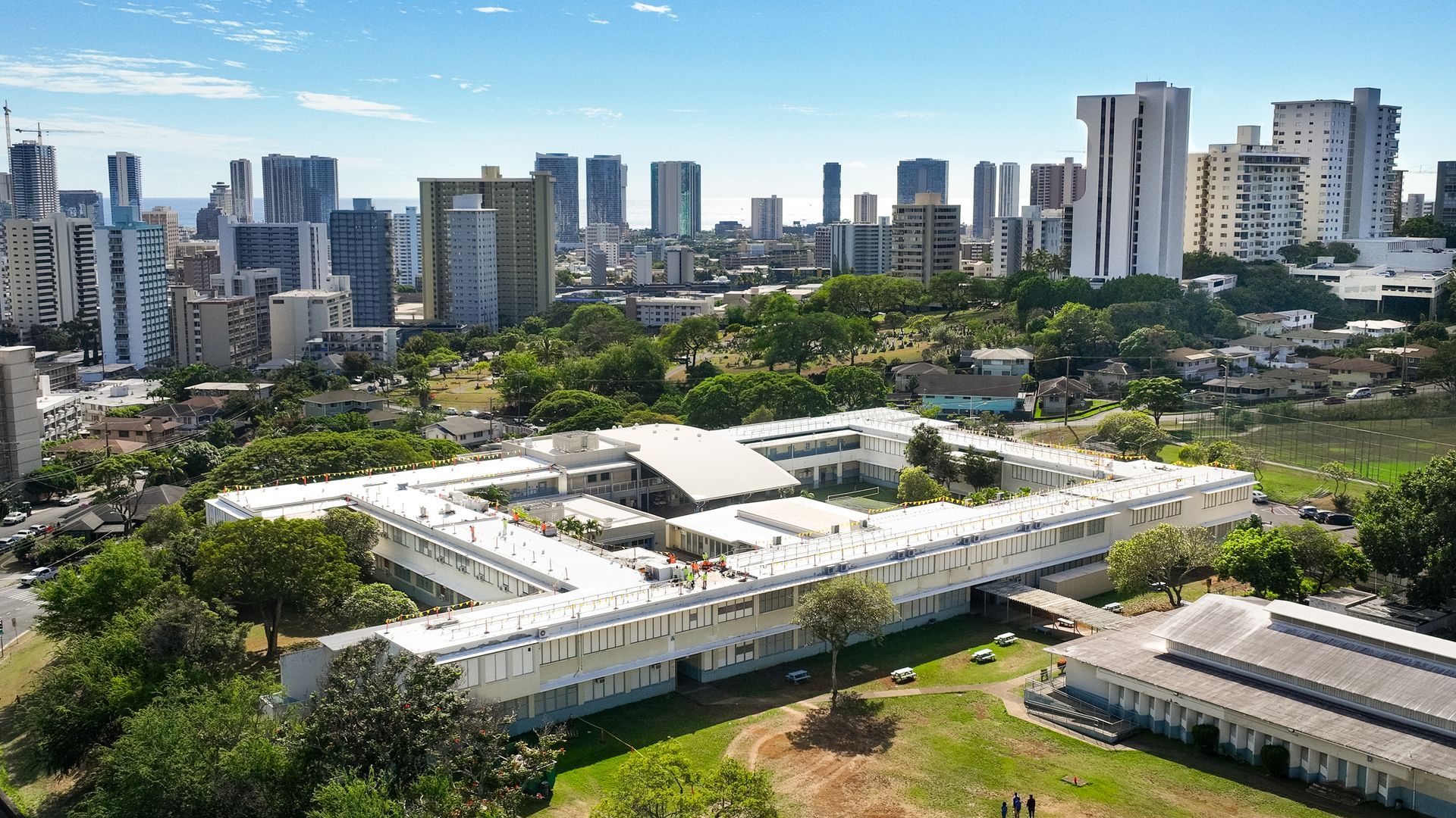 Aerial view of a white, multi-building structure with a city skyline in the background. Green trees and grass surround the buildings.