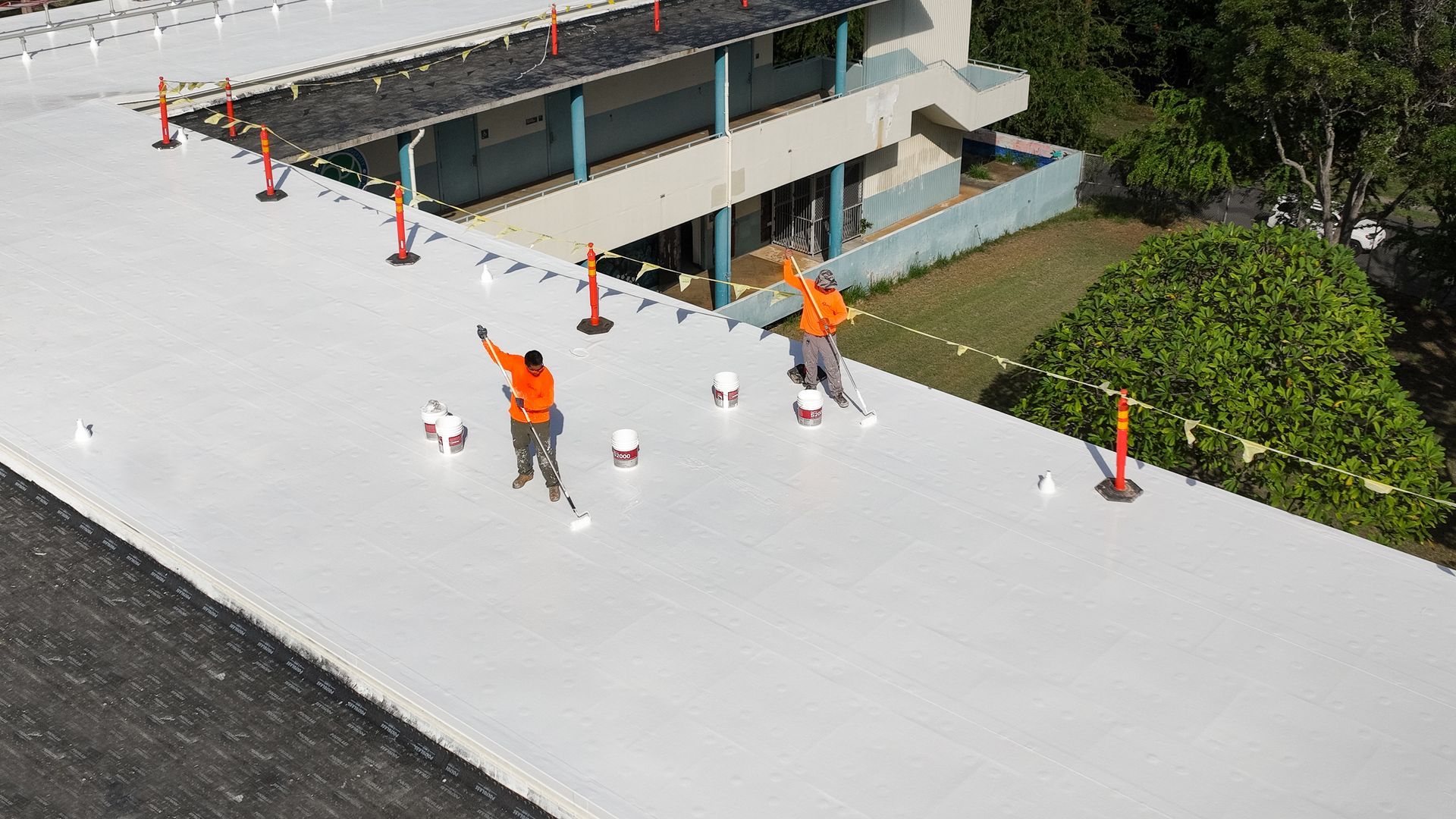 Workers in orange vests on a white roof, installing safety cones near a building, bright sunshine.