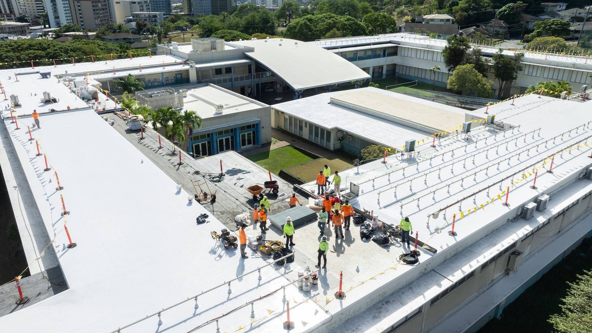 Construction workers on a flat white roof of a building with a central courtyard and surrounding trees.