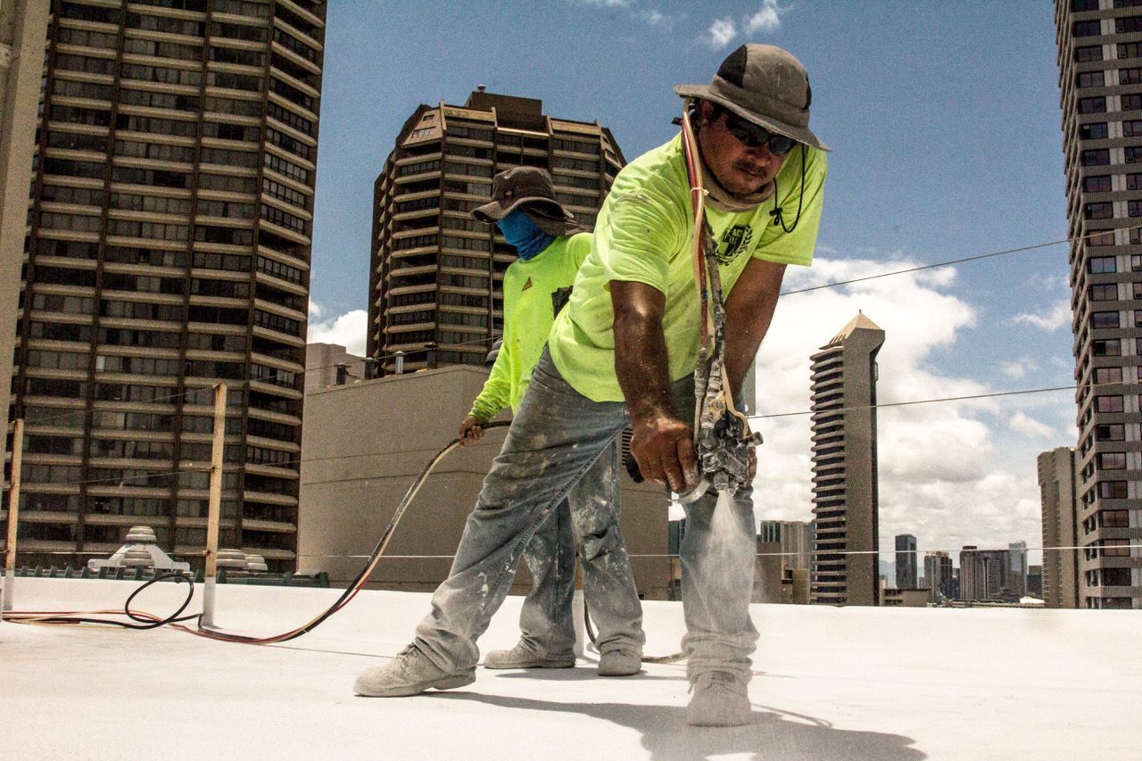 Two workers spray-paint a rooftop, wearing safety gear, amidst city skyscrapers.