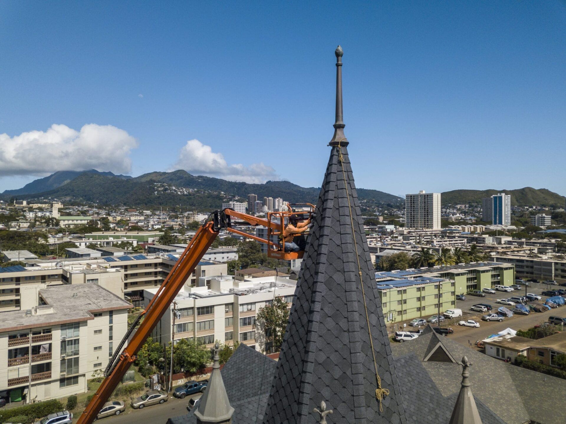 Orange lift truck near church spire with cityscape and mountains in background.