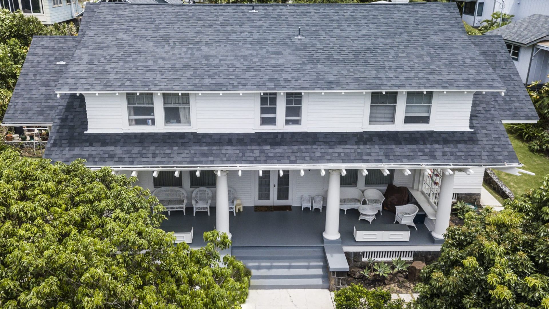 Two-story white house with a porch, surrounded by green trees and a grey roof.
