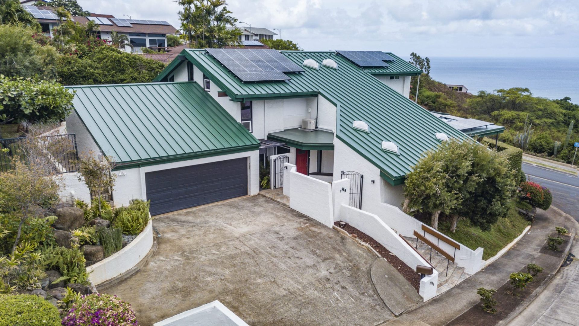 House with green roof and solar panels, gray garage, and driveway on a hillside overlooking the ocean.
