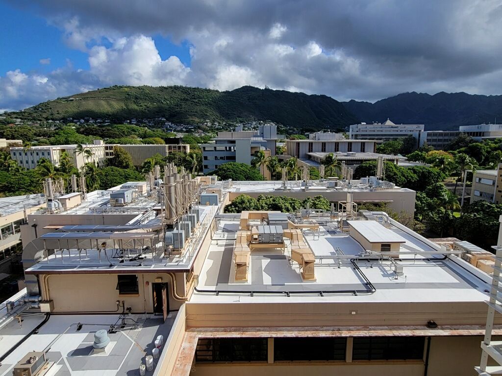 Rooftops of buildings with various equipment, trees, and mountains in the background under a cloudy sky.