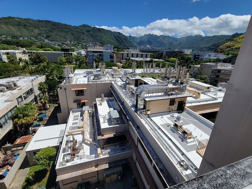 Rooftops of buildings with vents and equipment against a backdrop of green mountains and a blue sky.
