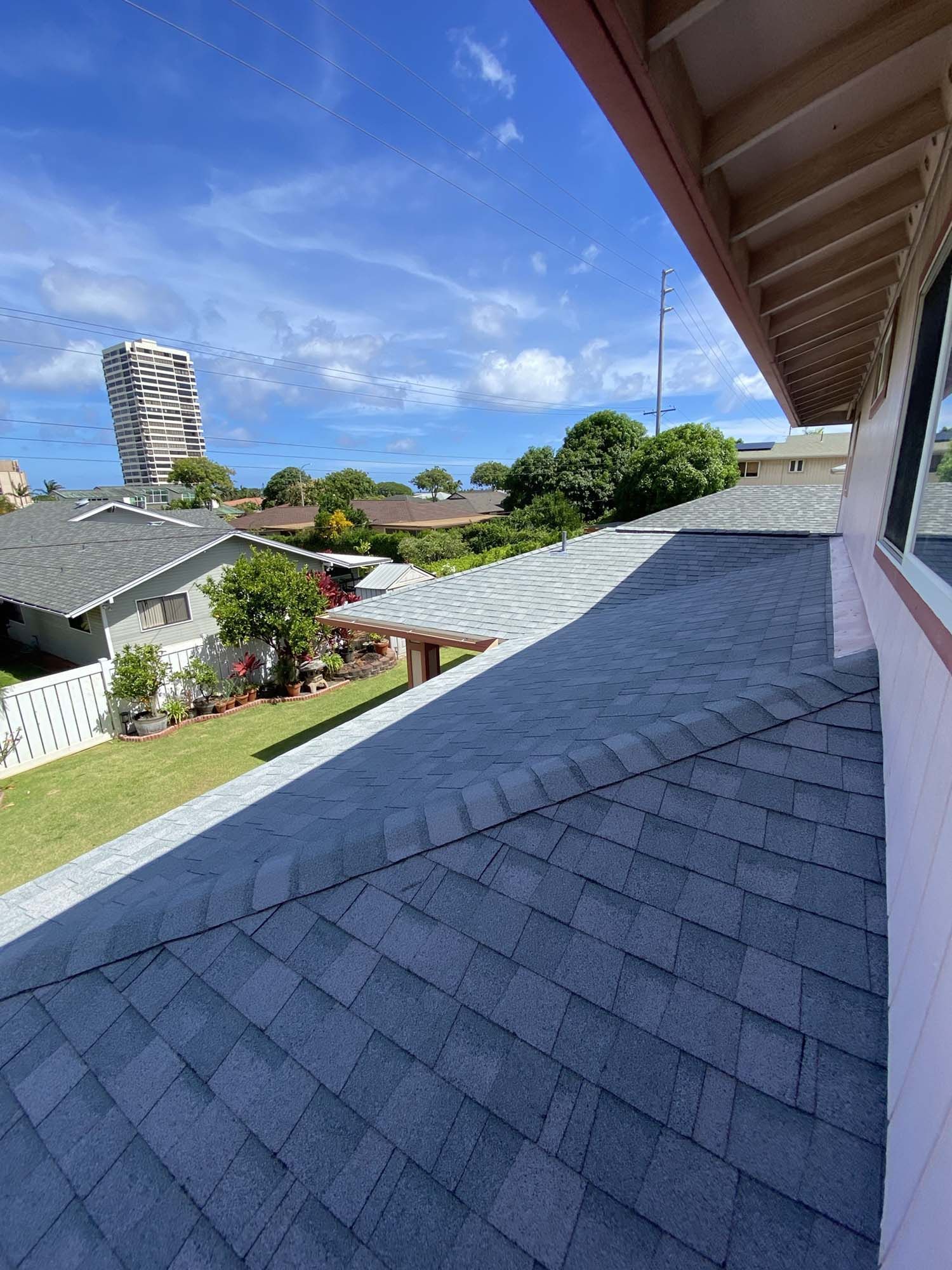 View from a roof: A gray shingled roof with a blue sky, buildings, and trees in the background.