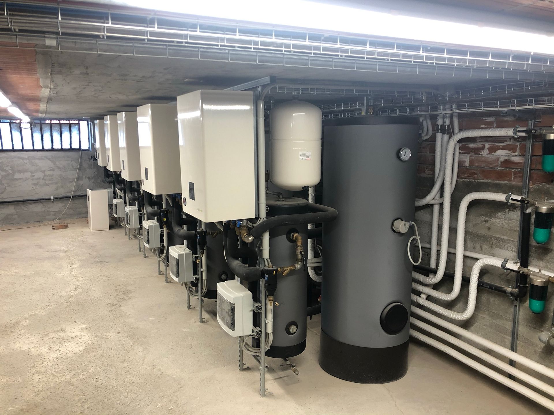 Fila di unità di riscaldamento industriali in un seminterrato. Apparecchiature bianche e grigie, tubi e un grande serbatoio d'acqua.