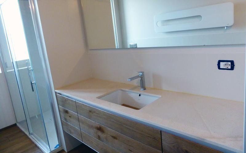 Bagno con piano di lavoro bianco, mobili in legno, lavandino, specchio e doccia con porta in vetro.