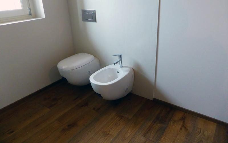 WC e bidet in un bagno con pavimento in legno scuro, pareti bianche e una piccola finestra.