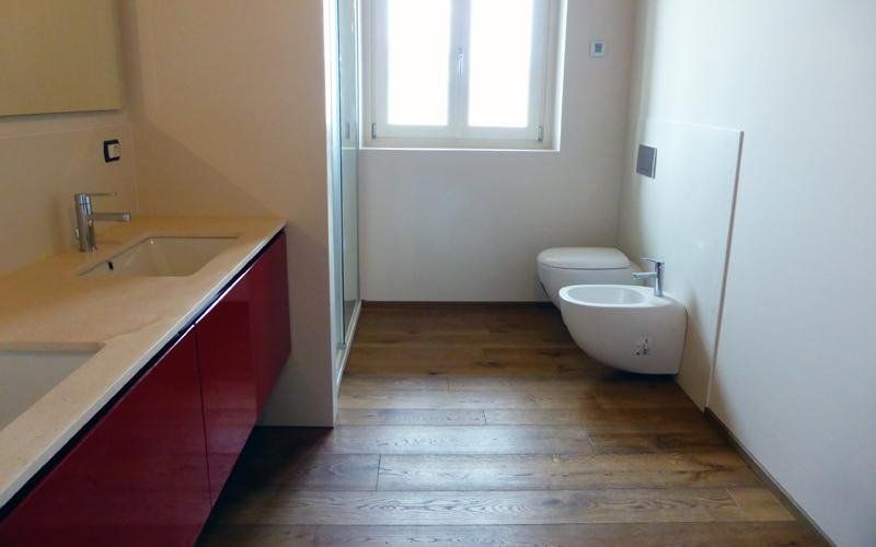 Bagno moderno con mobiletto rosso, lavabo, WC, bidet e pavimento in legno. Luce naturale e intensa proveniente dalla finestra.