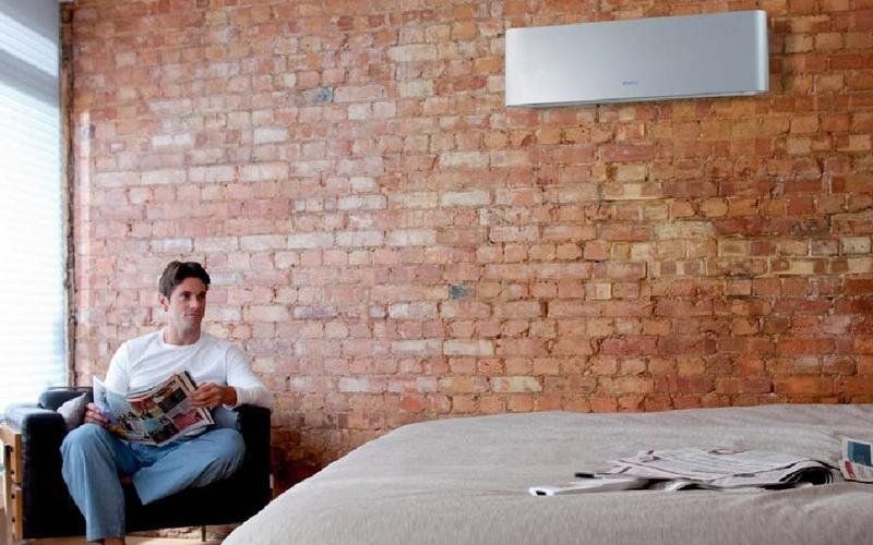 Uomo seduto su una sedia, che legge una rivista, in una stanza con un muro di mattoni e un condizionatore sopra il letto.