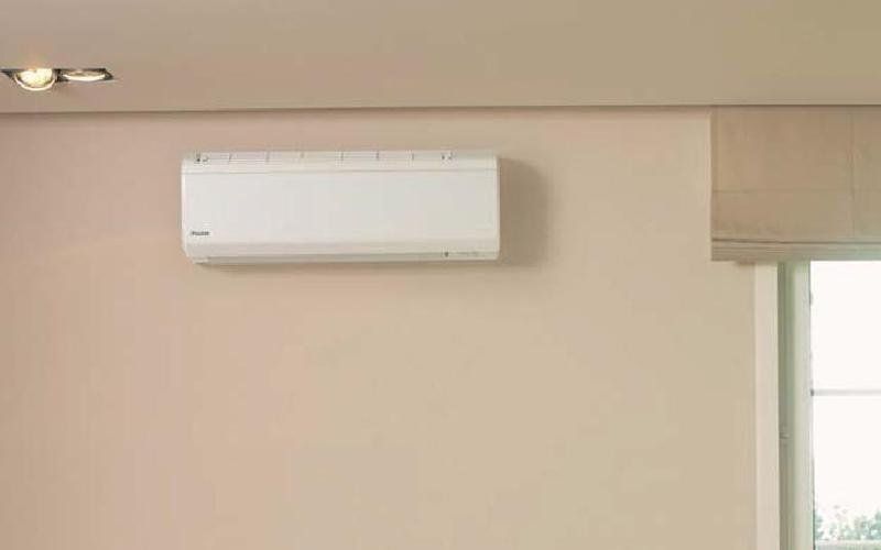 Condizionatore d'aria bianco montato a parete su una parete beige, accanto a una finestra con una tenda beige.