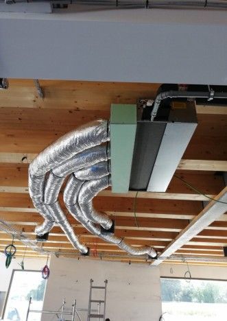 Installazione di condotti HVAC in un edificio. Condotti isolati in argento fuoriescono da un'unità verde e nera, fissata a un soffitto in legno.