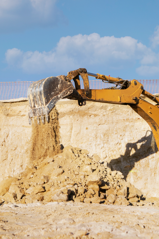 Escavadeira amarela despejando areia em um canteiro de obras; céu azul ao fundo.