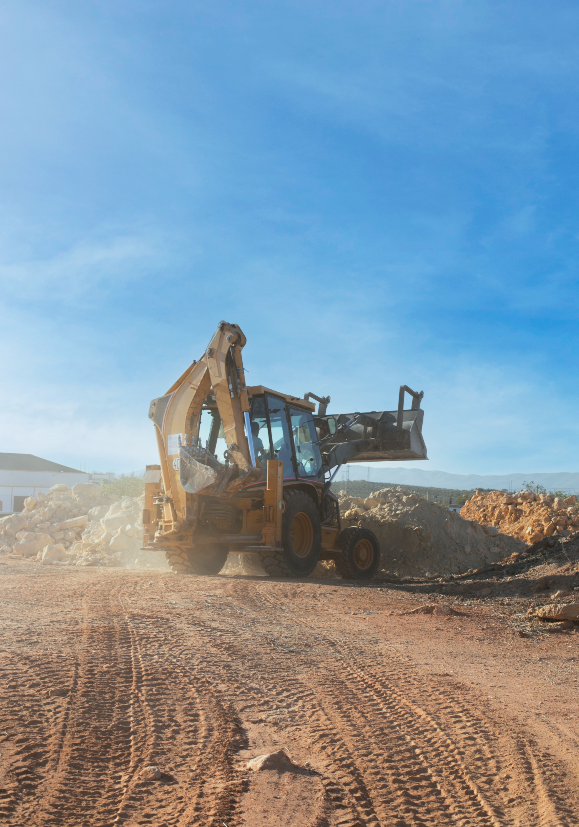 Retroescavadeira amarela operando em uma estrada de terra, levantando pedras sob um céu azul.