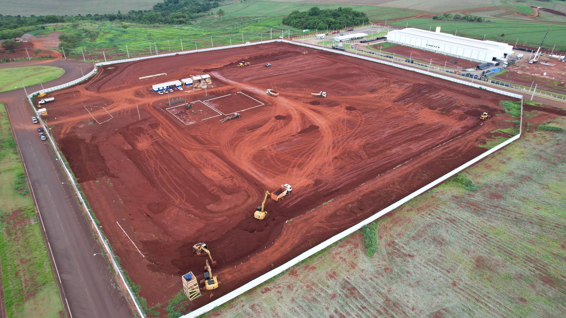 Vista aérea de um canteiro de obras com máquinas de terraplenagem, solo vermelho e um prédio com telhado branco.