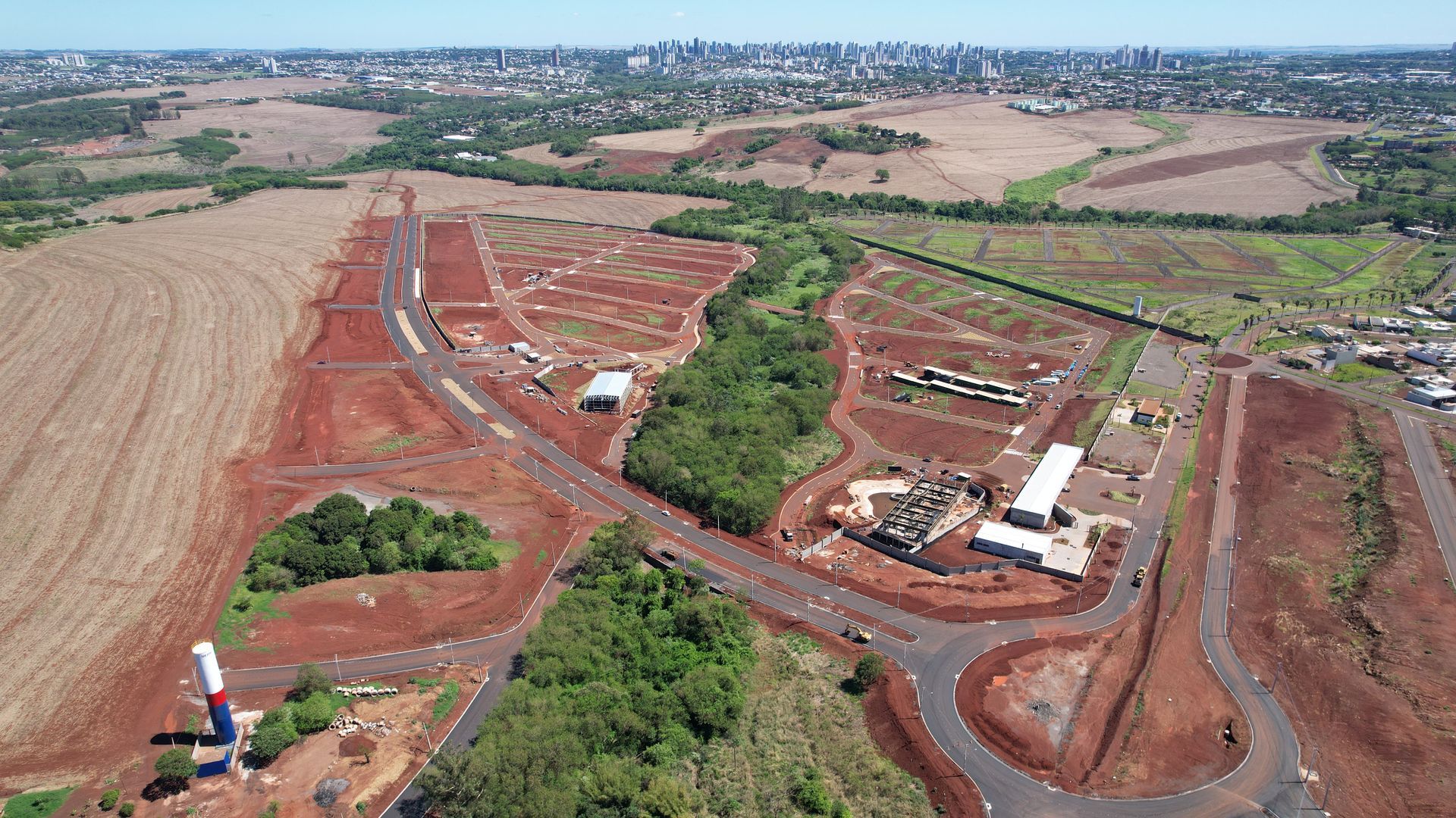 Vista aérea de um canteiro de obras com terreno limpo, terra vermelha, edifícios e o horizonte da cidade ao longe.
