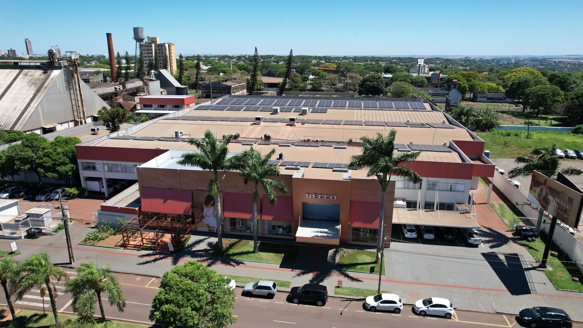 Vista aérea de um grande edifício com detalhes em vermelho, palmeiras e carros estacionados, sob um céu limpo.