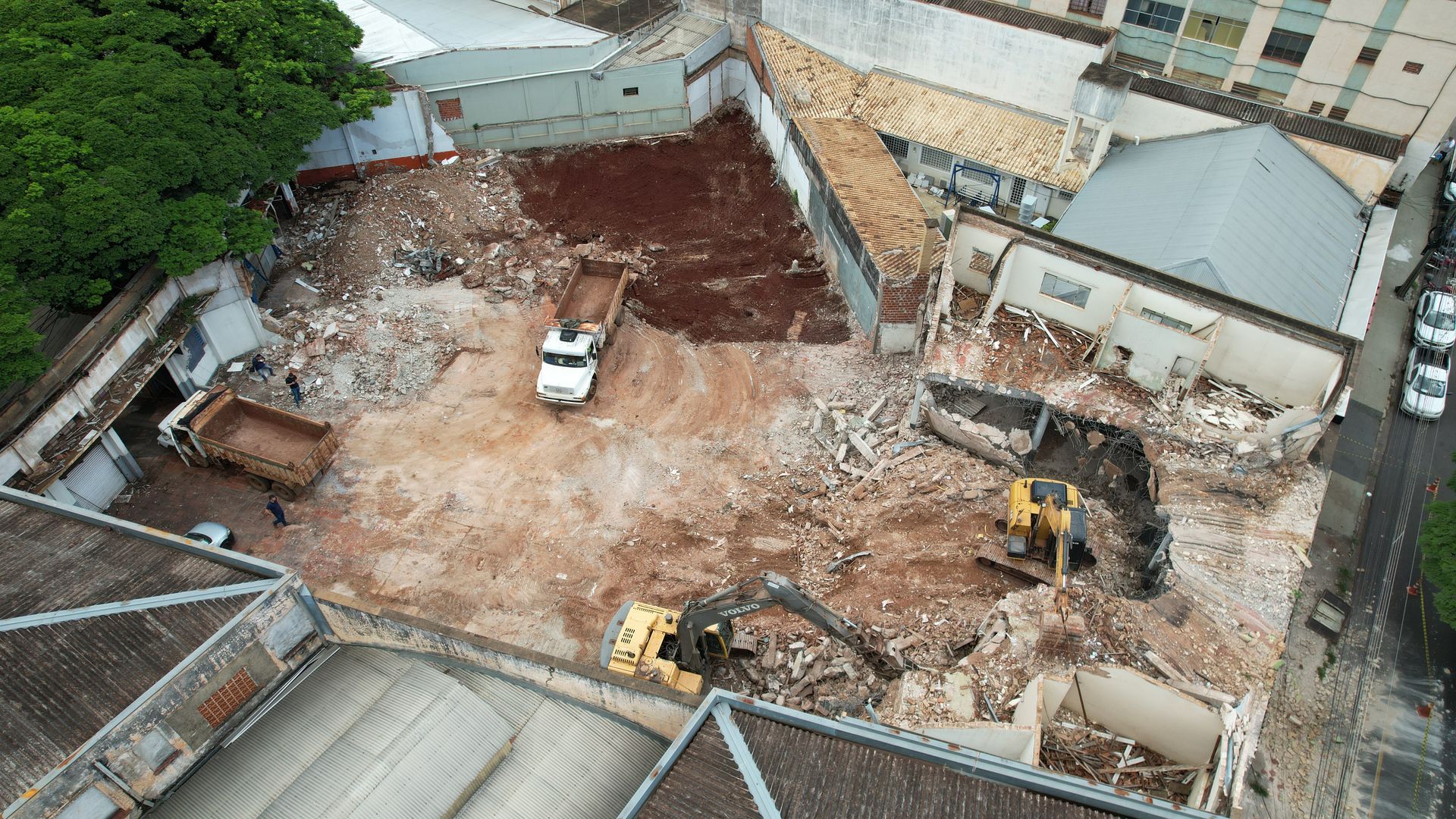 Vista aérea de um local de demolição com equipamentos de construção, entulho e caminhões cercados por edifícios.