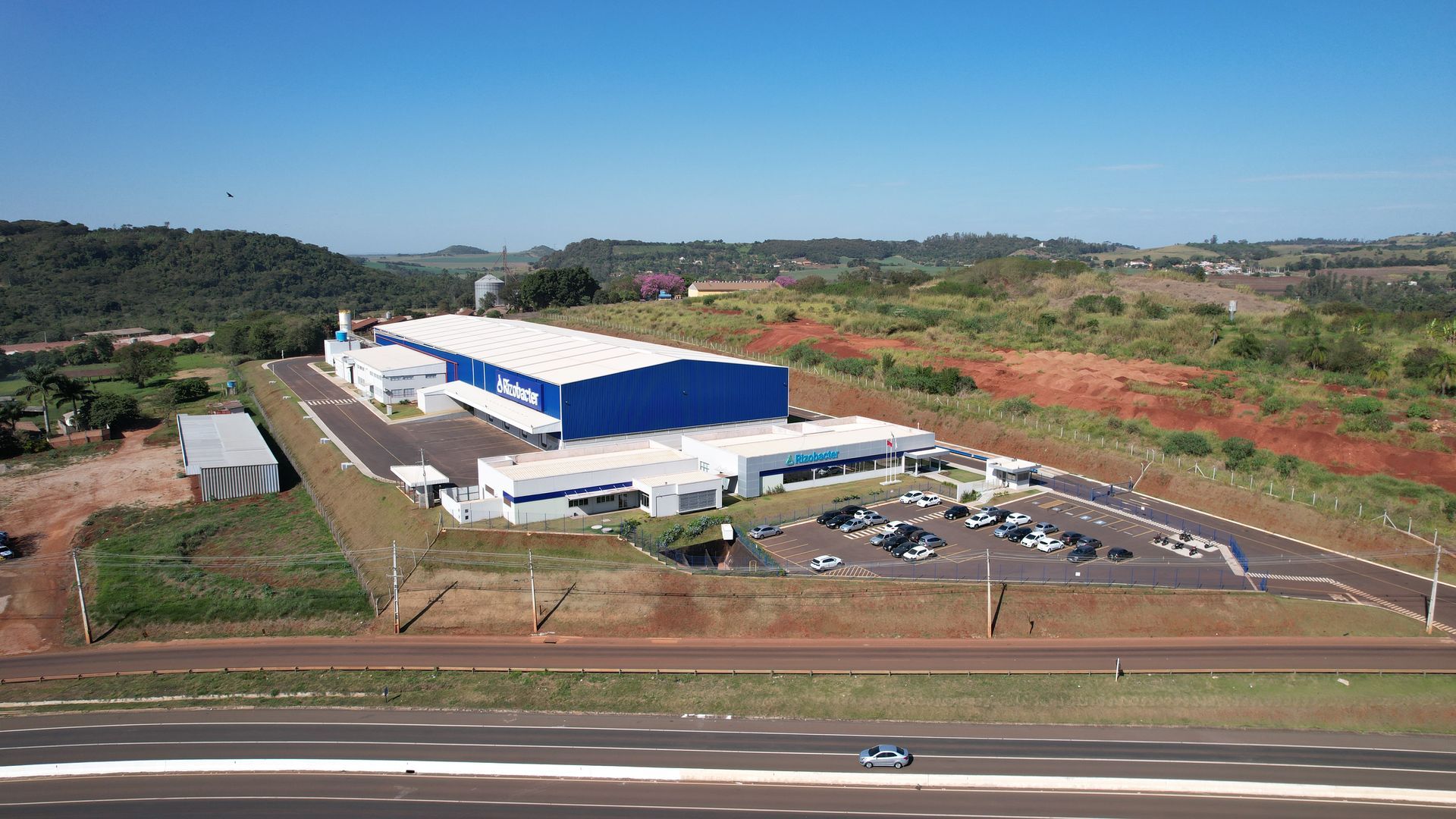 Grande edifício industrial azul e branco com estacionamento, situado em uma colina com vegetação.