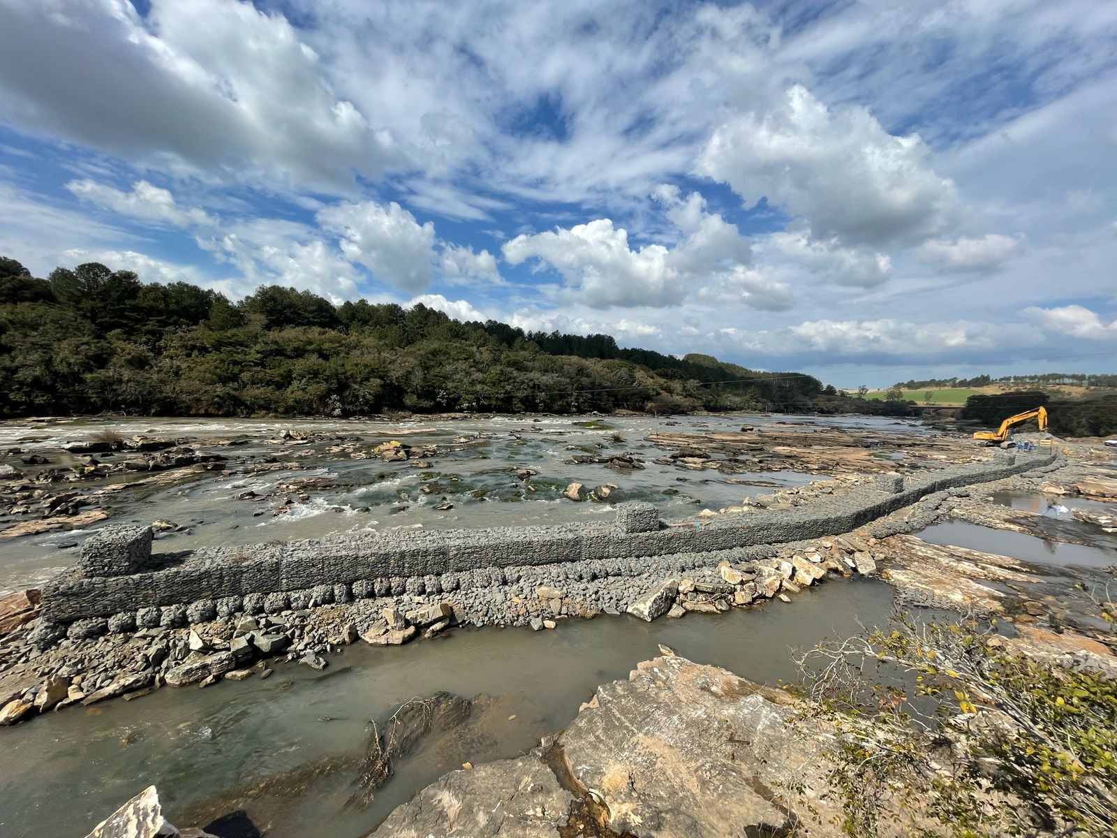 Canteiro de obras perto da água, com céu nublado e uma escavadeira ao longe.