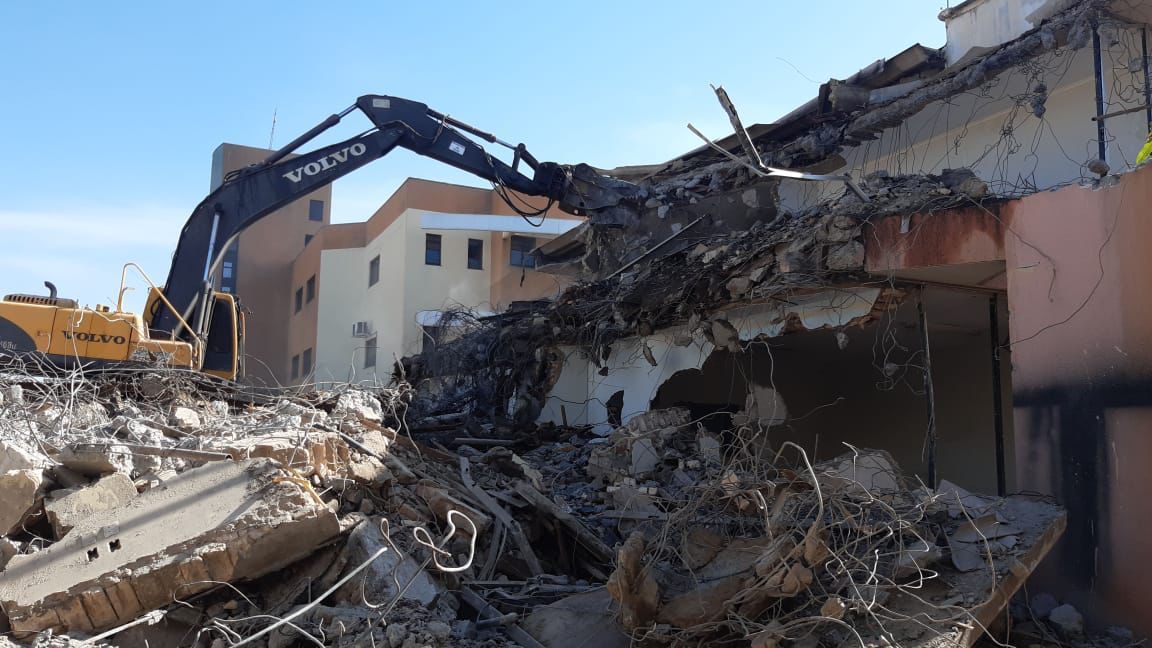 Uma escavadeira demoli um prédio, pilhas de entulho visíveis, céu azul.