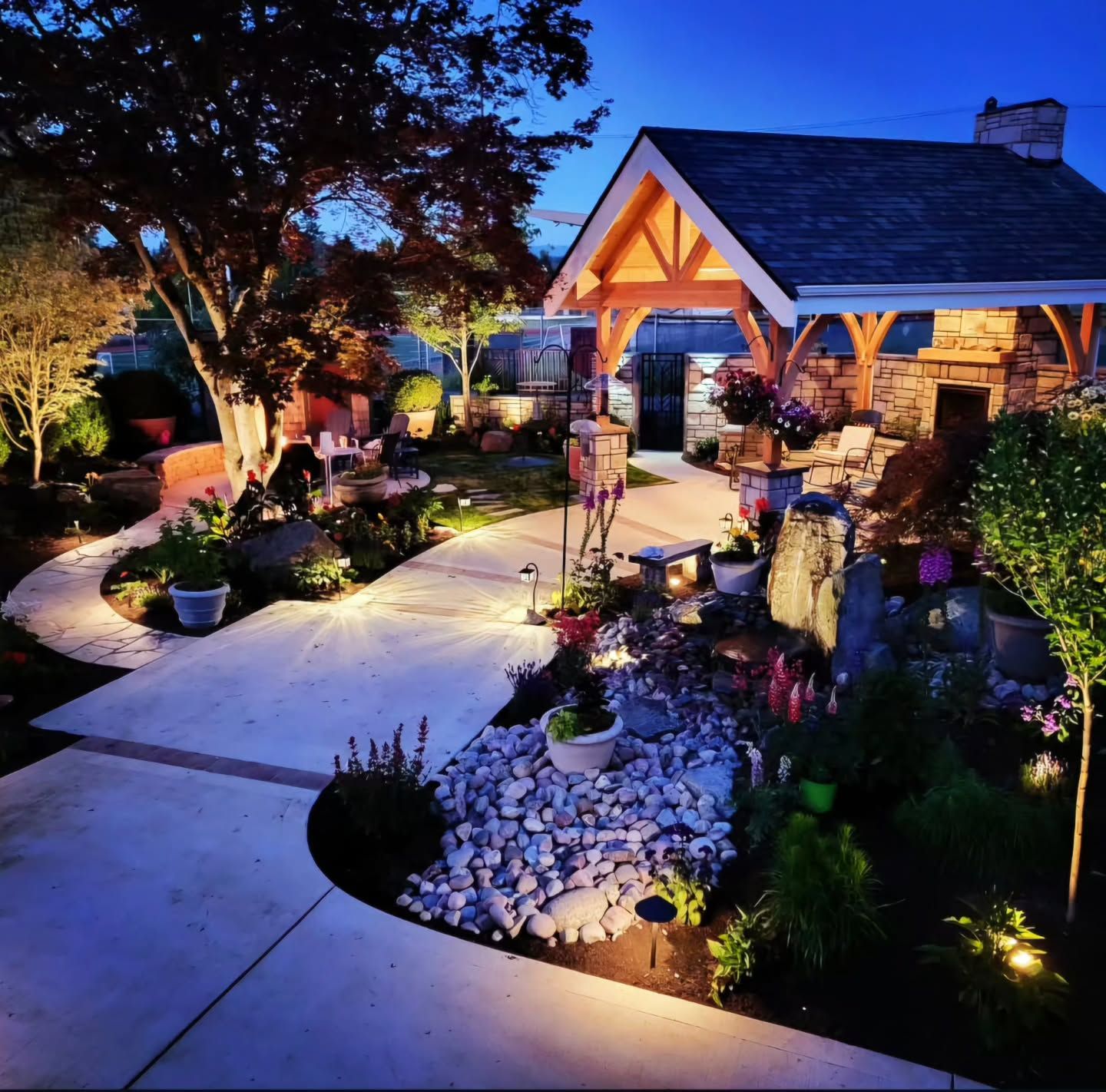 Lit backyard with a stone and wood pavilion, a concrete path, and landscaping at dusk.