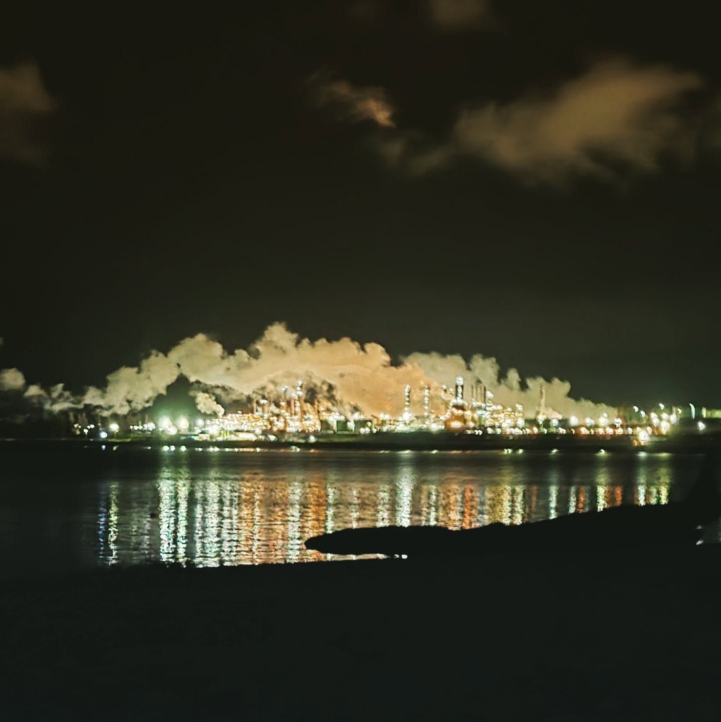 Night scene of a lit industrial complex emitting smoke, reflecting on the water below.
