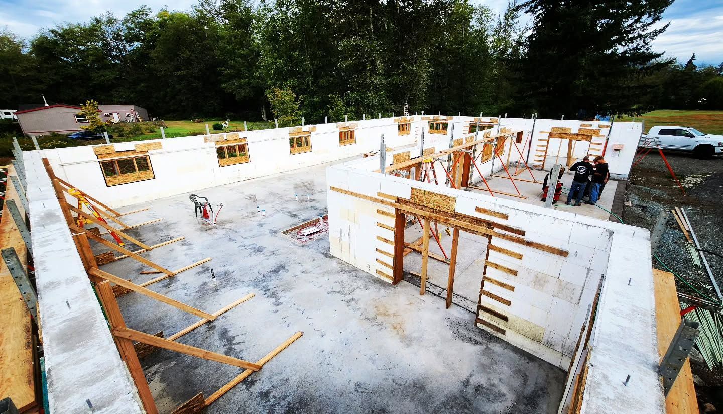 Foundation of a building under construction; white walls with wooden supports, workers, and trees in the background.