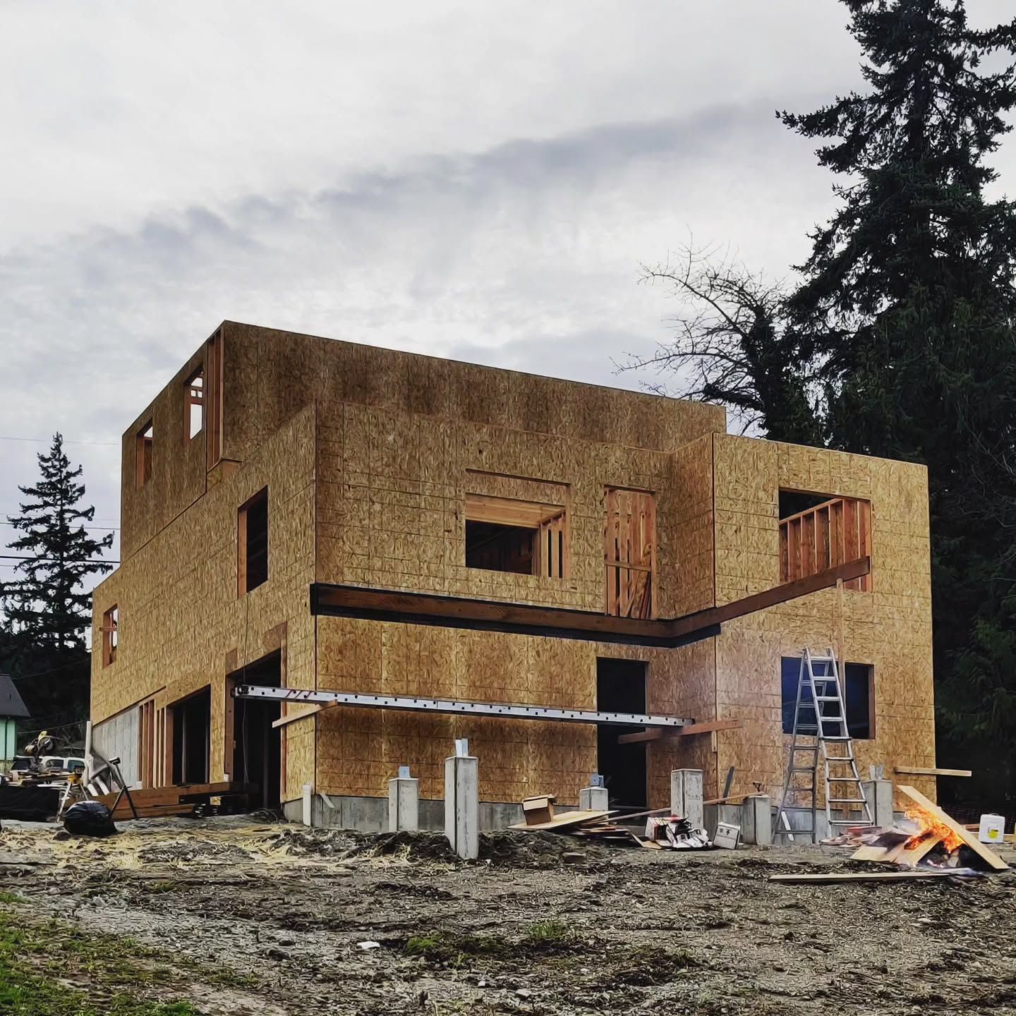 Two-story house under construction, framed with wood, concrete foundation, cloudy day, trees in the background.