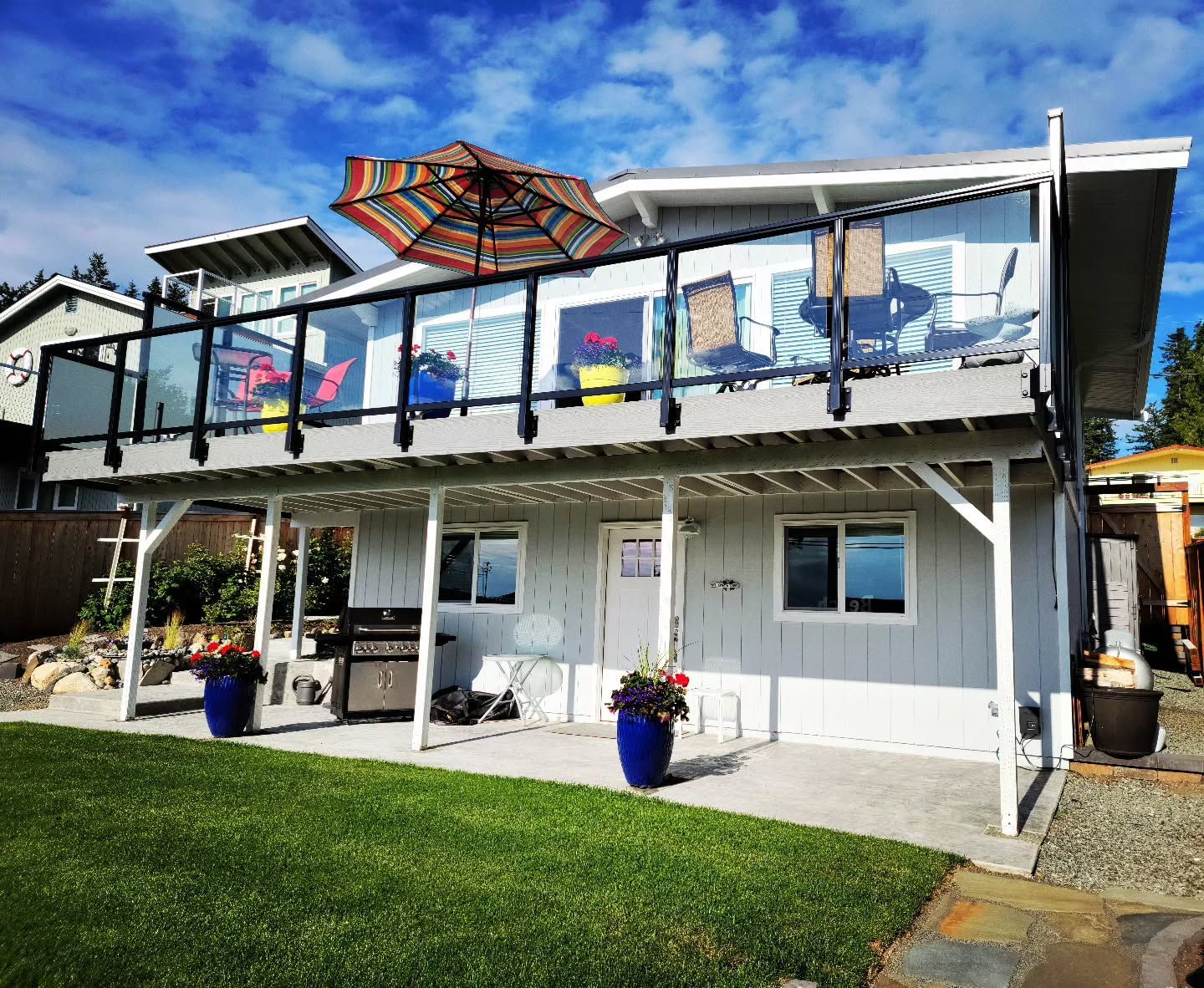 Two-story beach house with glass-railed deck, umbrella, chairs. Gray exterior, blue pots, green grass, sunny day.