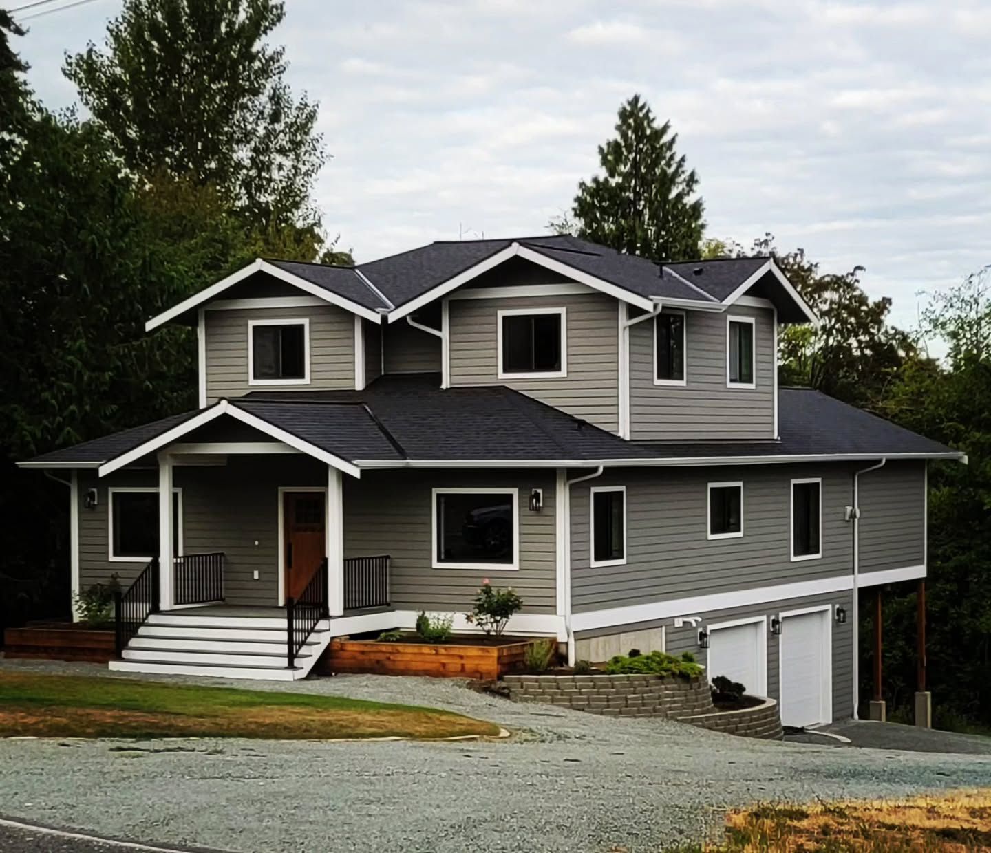 Gray two-story house with a dark roof, garage, and front porch, set on a slight slope; cloudy sky backdrop.