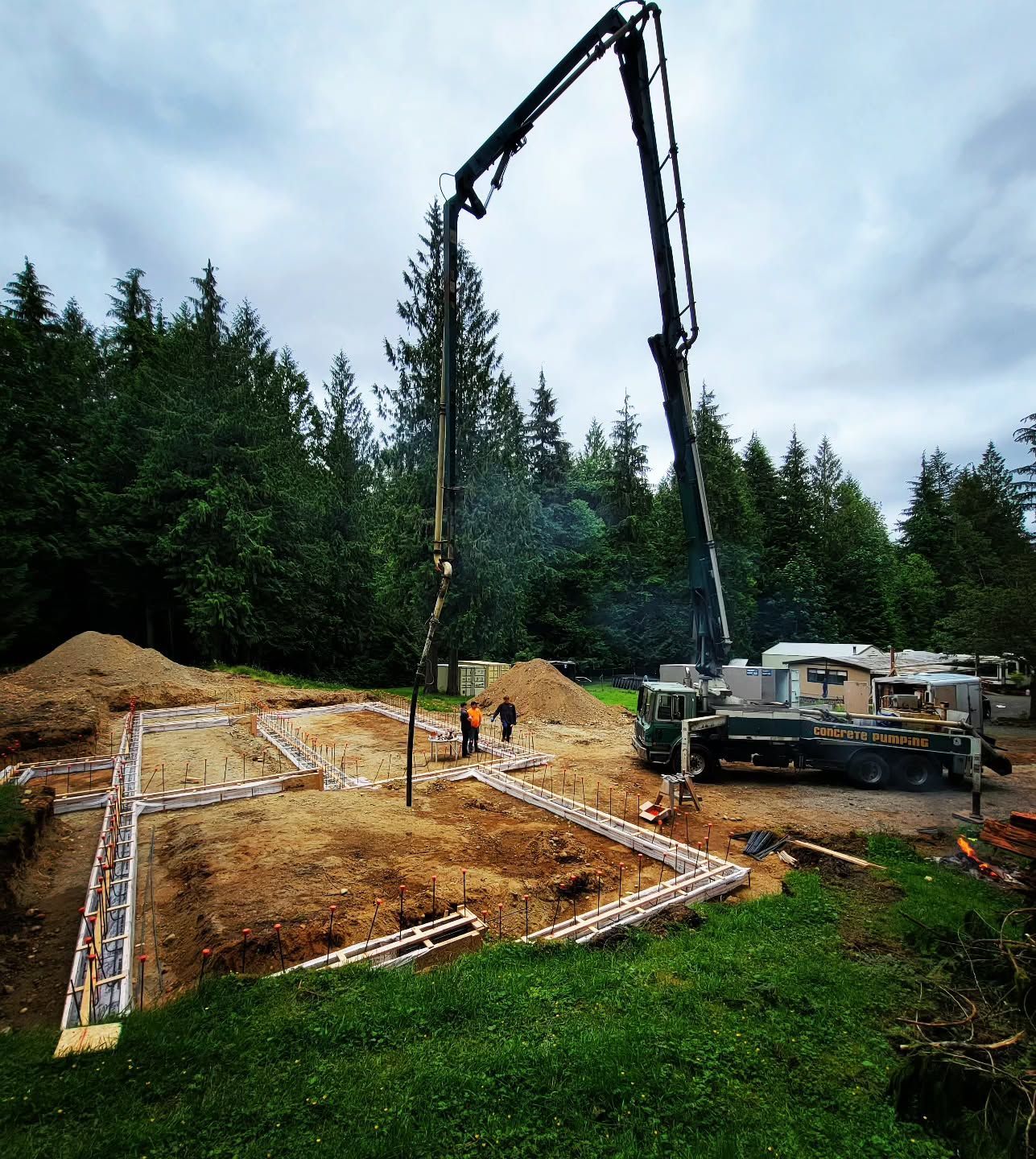 Construction site with a concrete pump truck pouring cement into foundation forms in a wooded area.