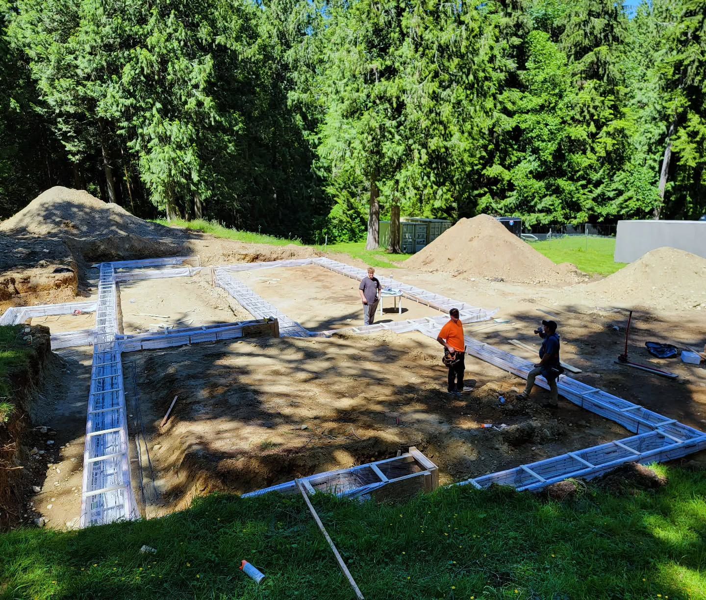 Construction site with foundation forms in place; three workers on site. Green trees in the background.
