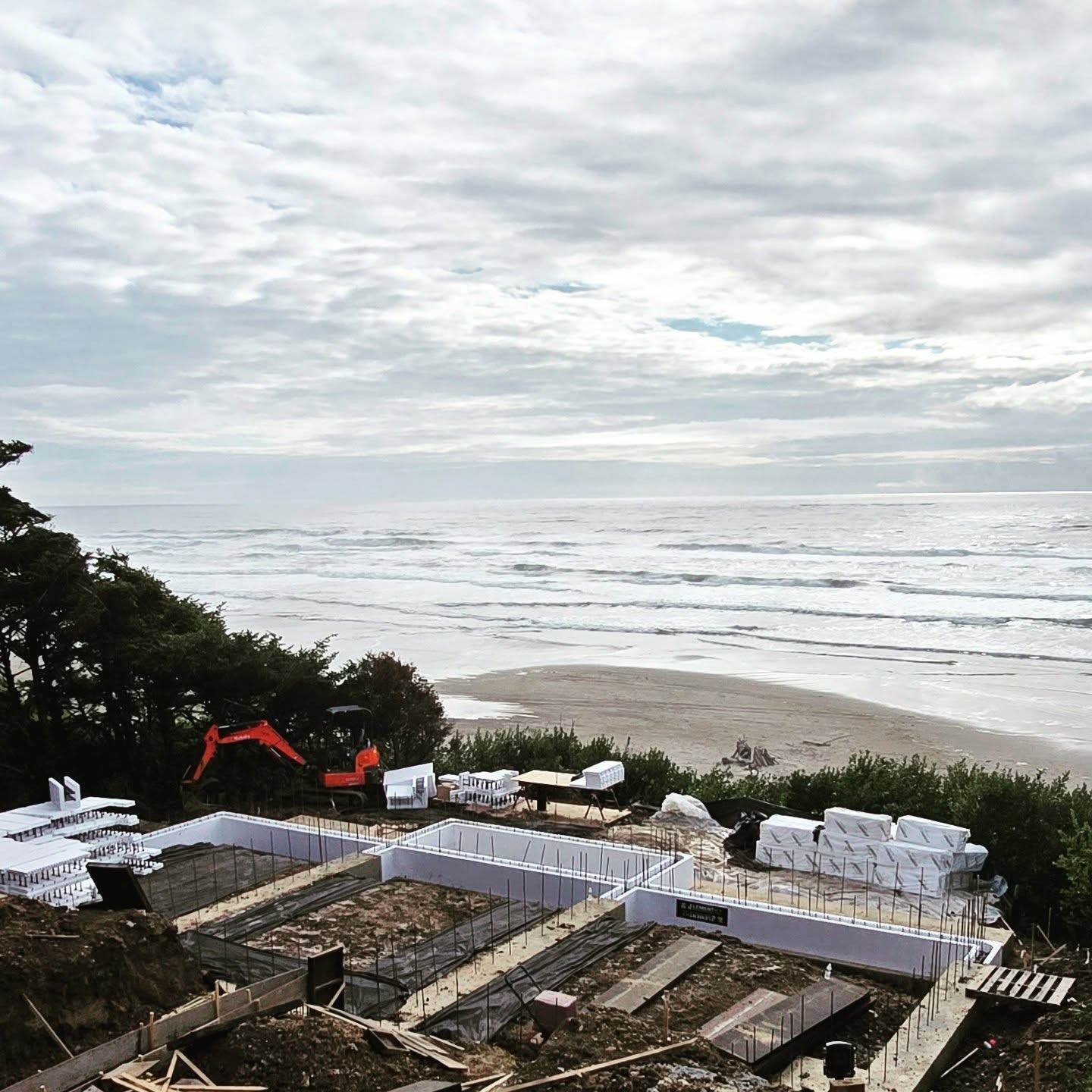 Construction site overlooking ocean: foundation being built, excavator, cloudy sky, sandy beach.