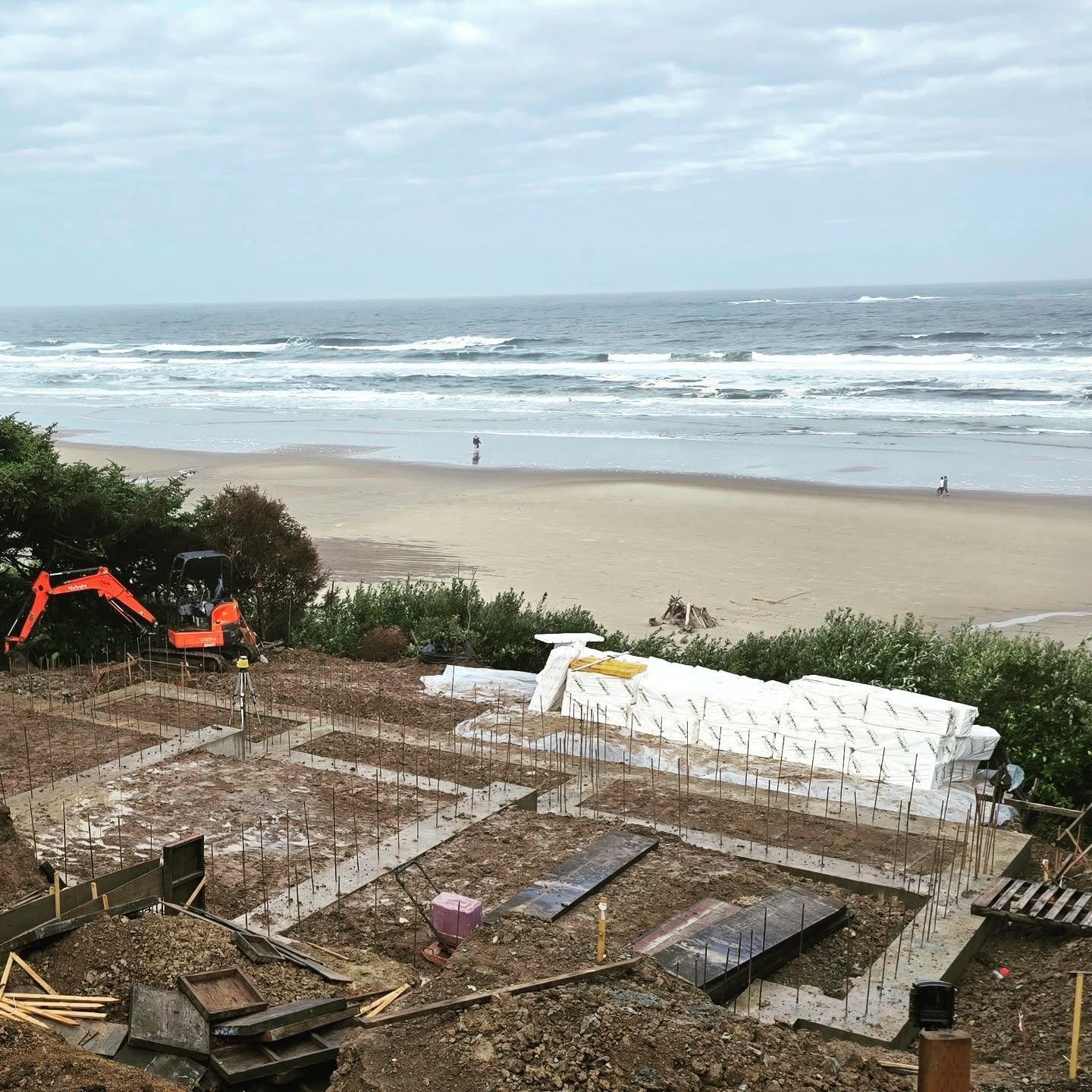 Construction site with ocean view: dirt foundation with rebar, excavator, beach, and waves.