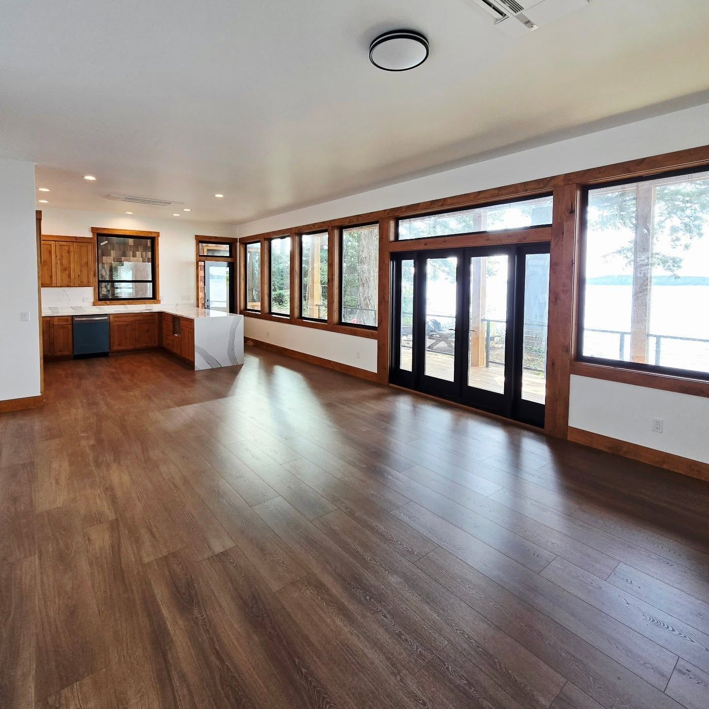 Spacious living room with dark wood floors, kitchen, and large windows overlooking a lake.