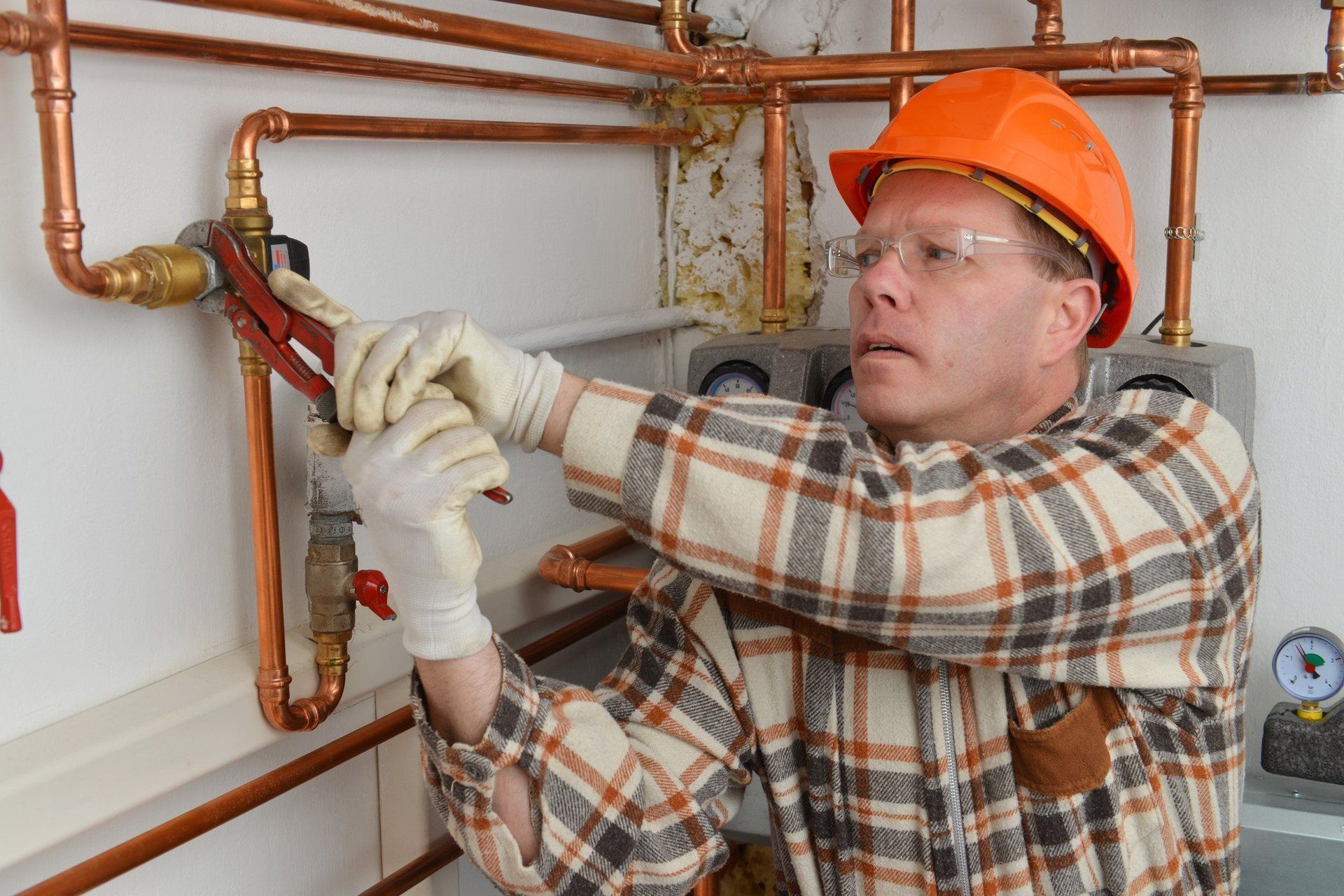 A person in a hard hat and plaid shirt uses a wrench to tighten a valve on a copper pipe system.