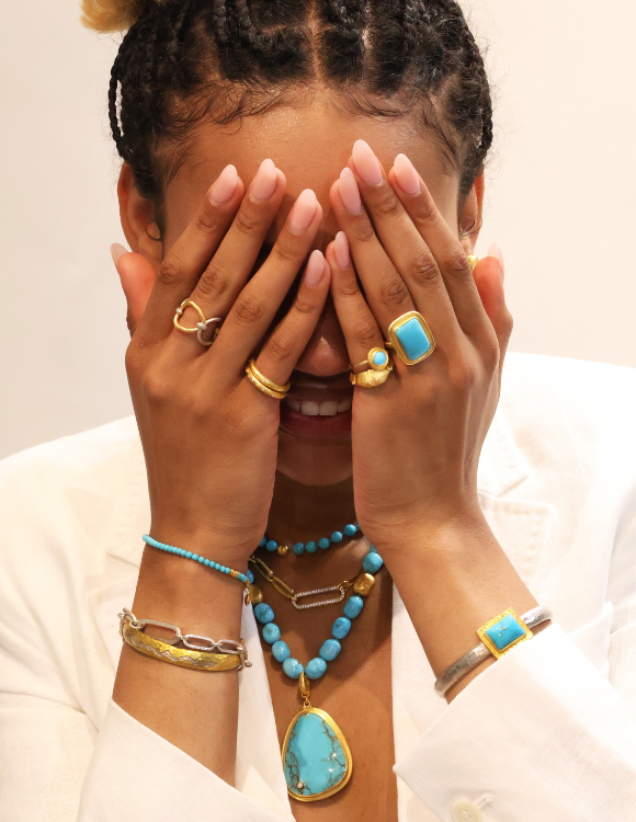 Person with hands covering face, showcasing turquoise jewelry, wearing a white blazer, indoors.