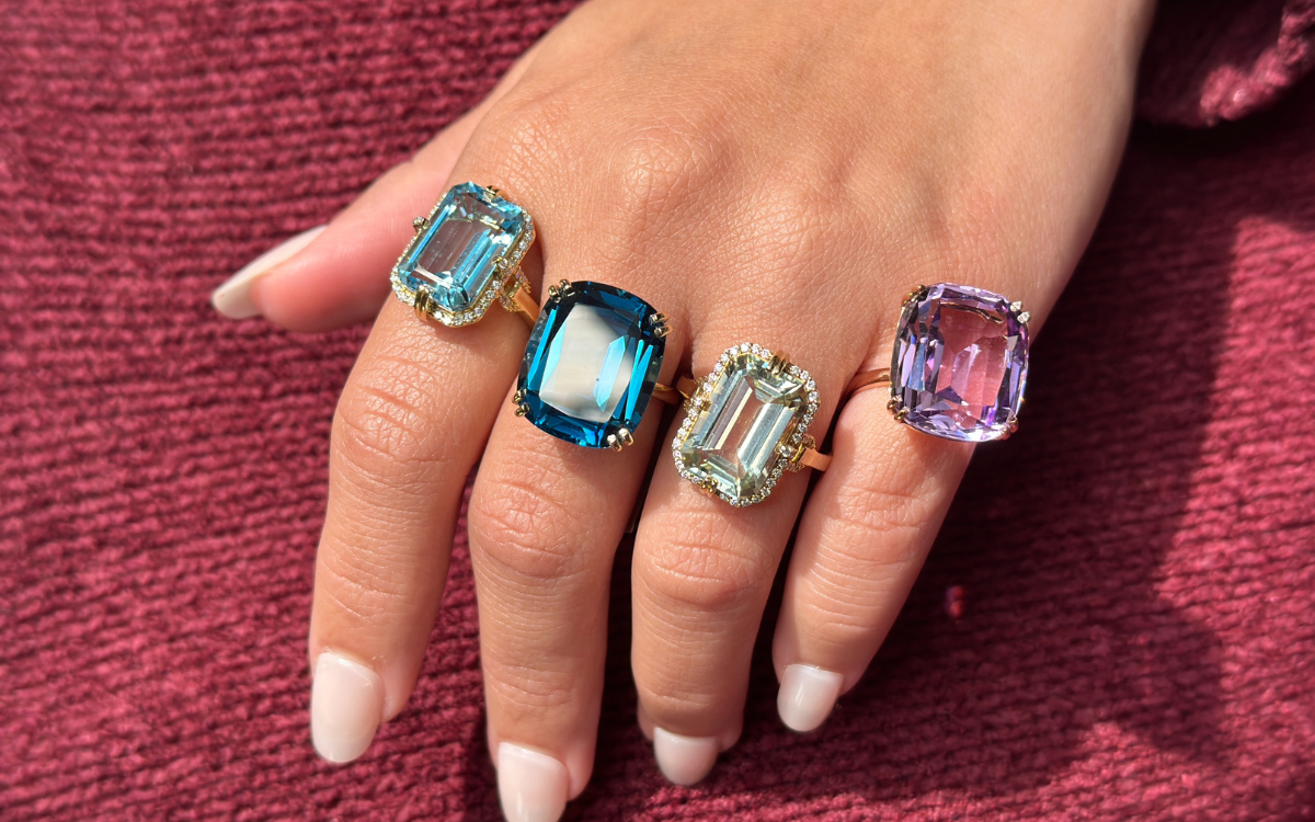 Hand wearing four gold rings with large colorful gemstones: blue, teal, green, purple.