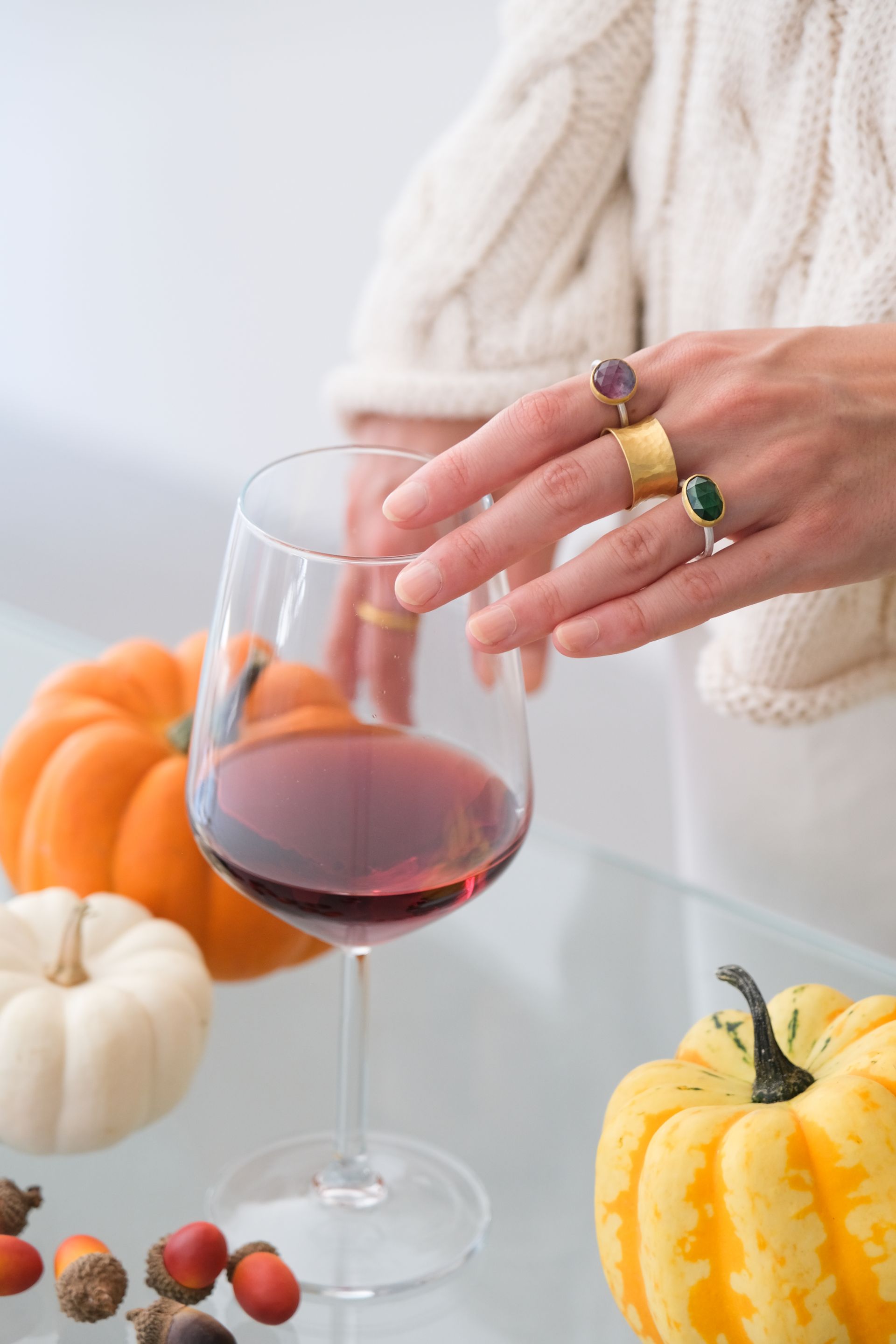 Hand with rings touches a glass of red wine, surrounded by gourds and acorns.