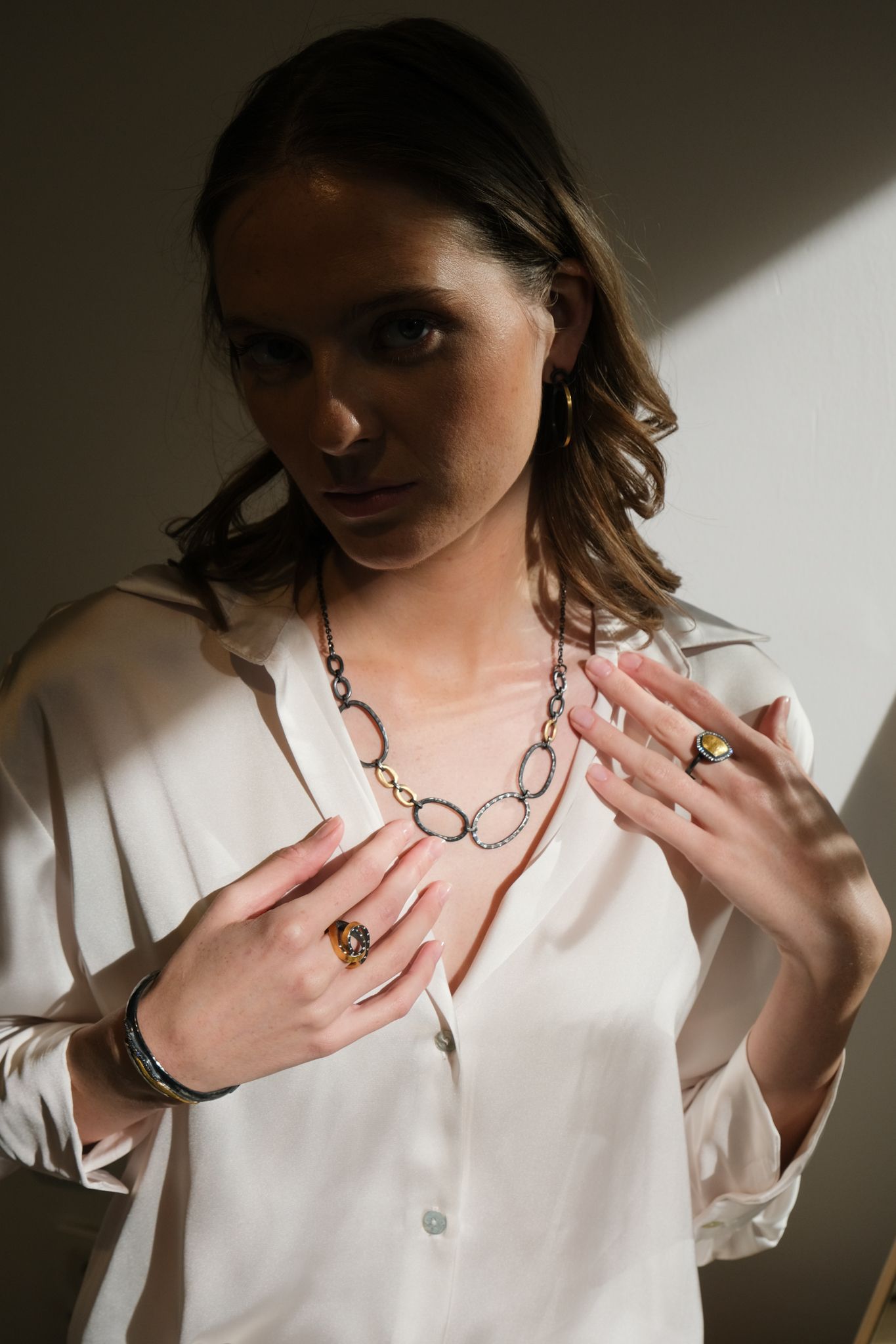 Woman wearing jewelry, white shirt, hands near chest, lit by sunlight.