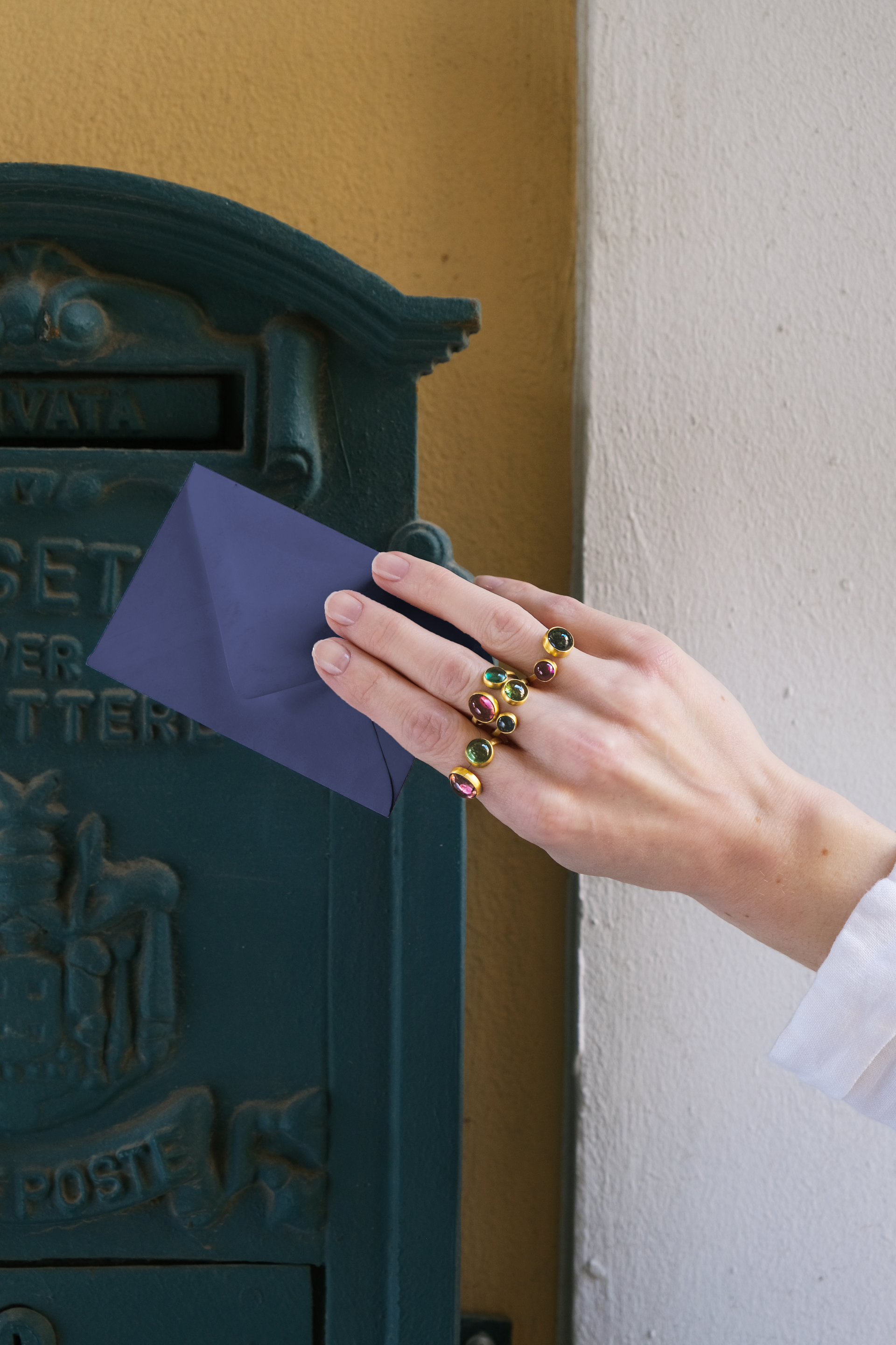 Hand placing a blue envelope into a dark green mailbox. Rings adorn the fingers.