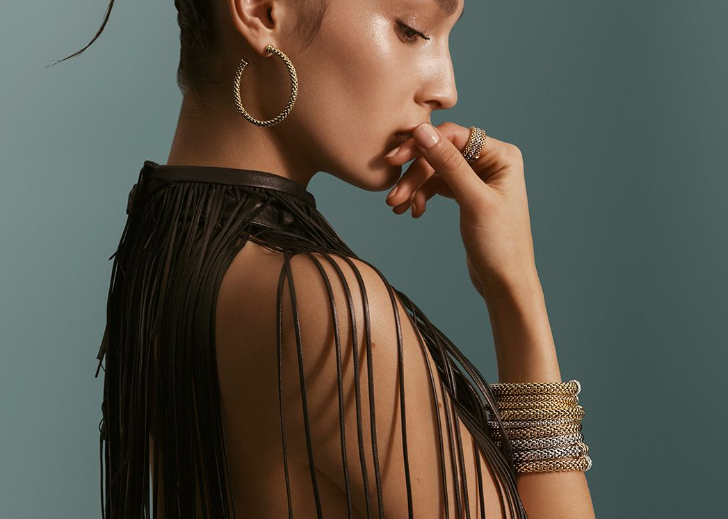 Woman in dark fringed top, wearing gold jewelry, hand near mouth, teal background.