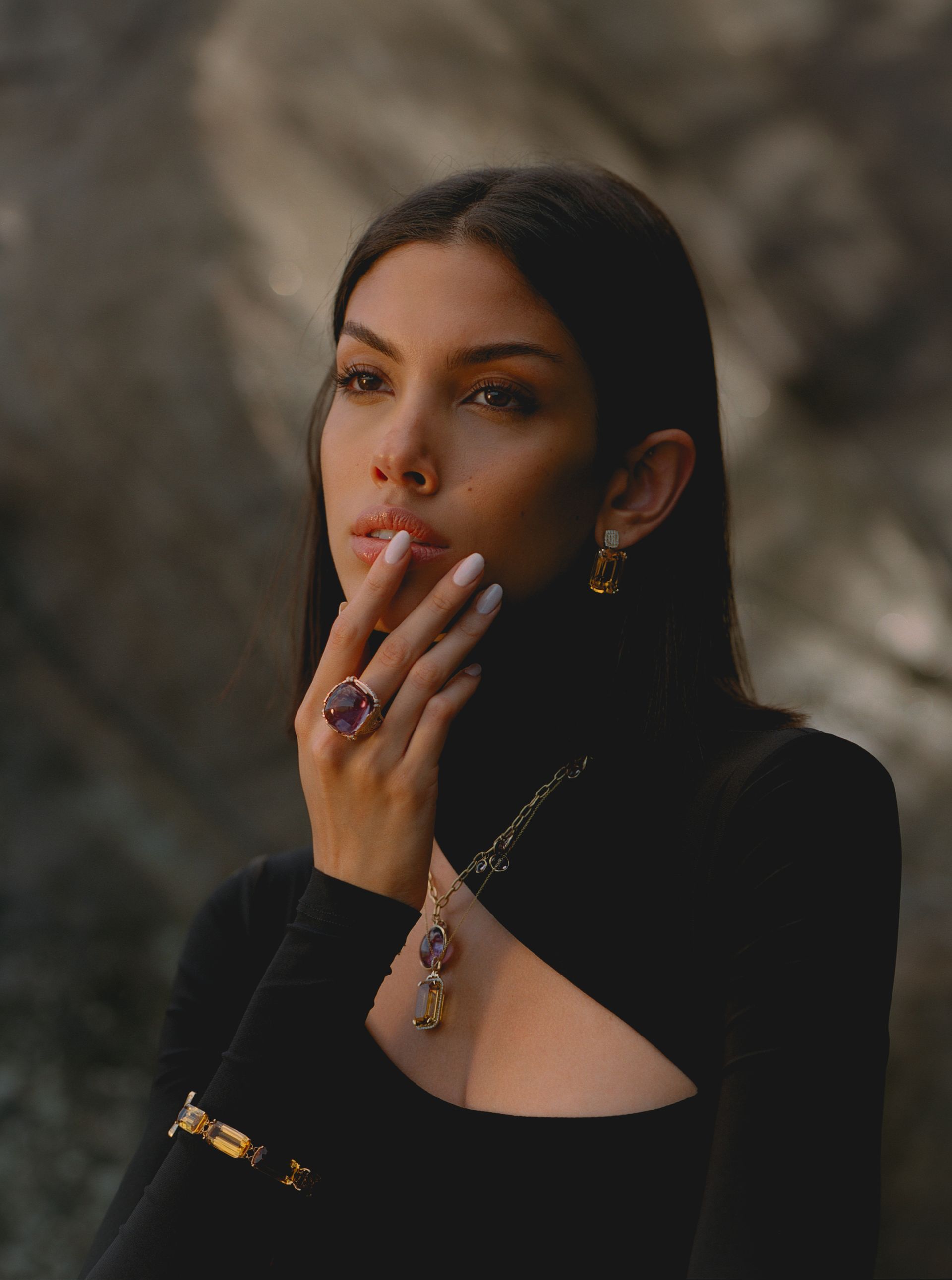 Woman in black turtleneck with jewelry, hand to face, outdoors.