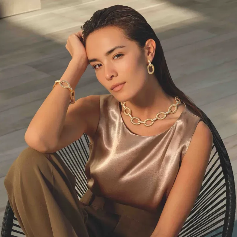 Woman wearing gold jewelry, seated in a chair, wearing a satin top and trousers.