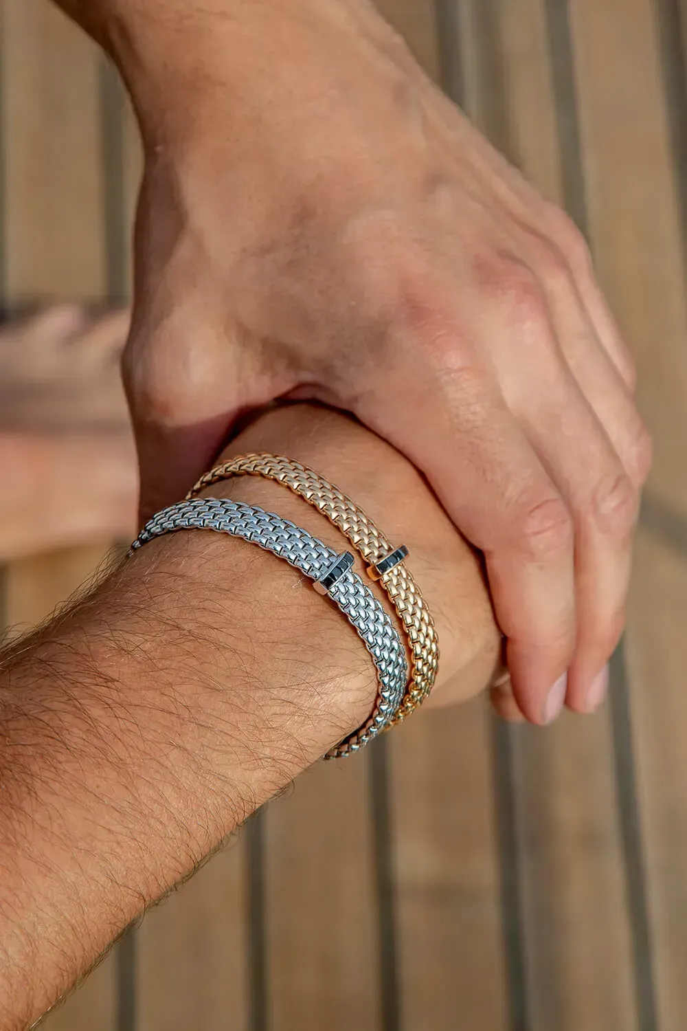 Two arms, one holding the other, each wearing a silver and gold woven bracelet.