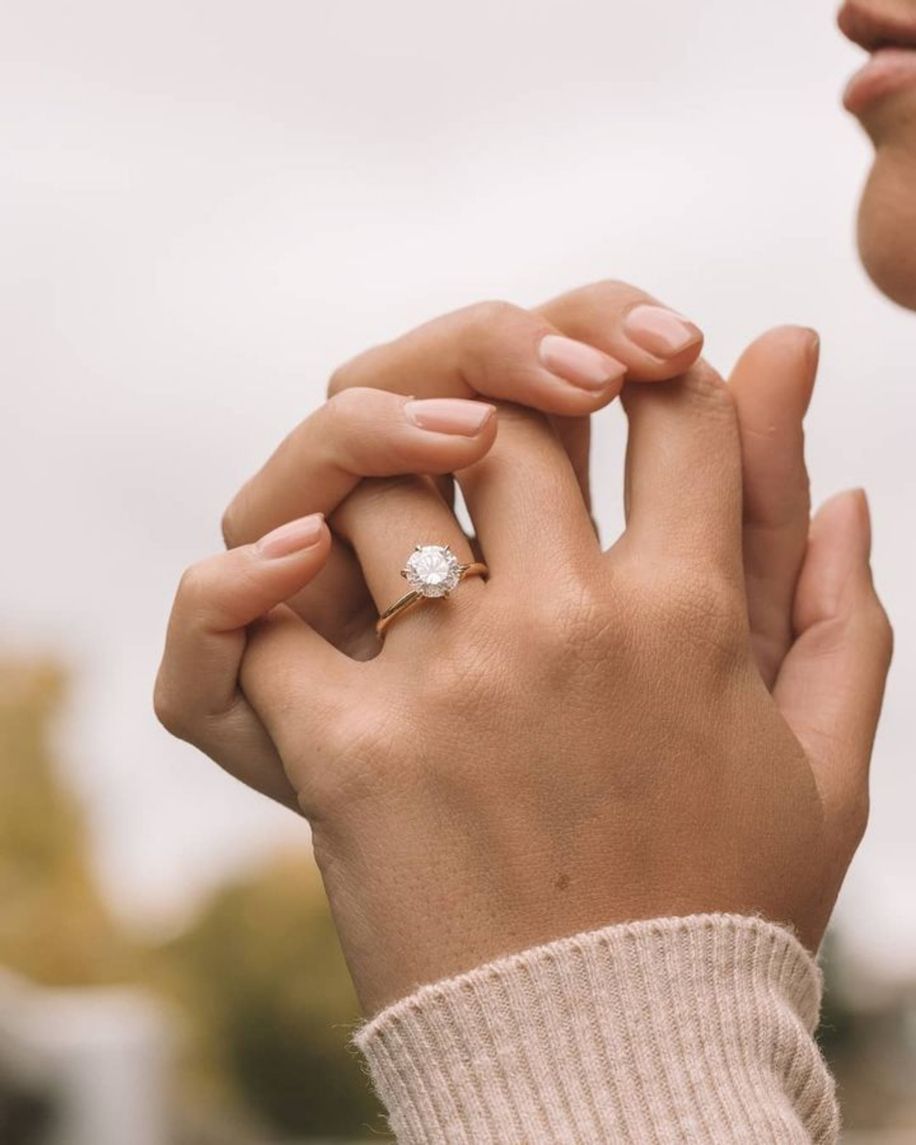 A person's hands holding, one with a gold ring and large clear stone. Pink sweater and blurred background.