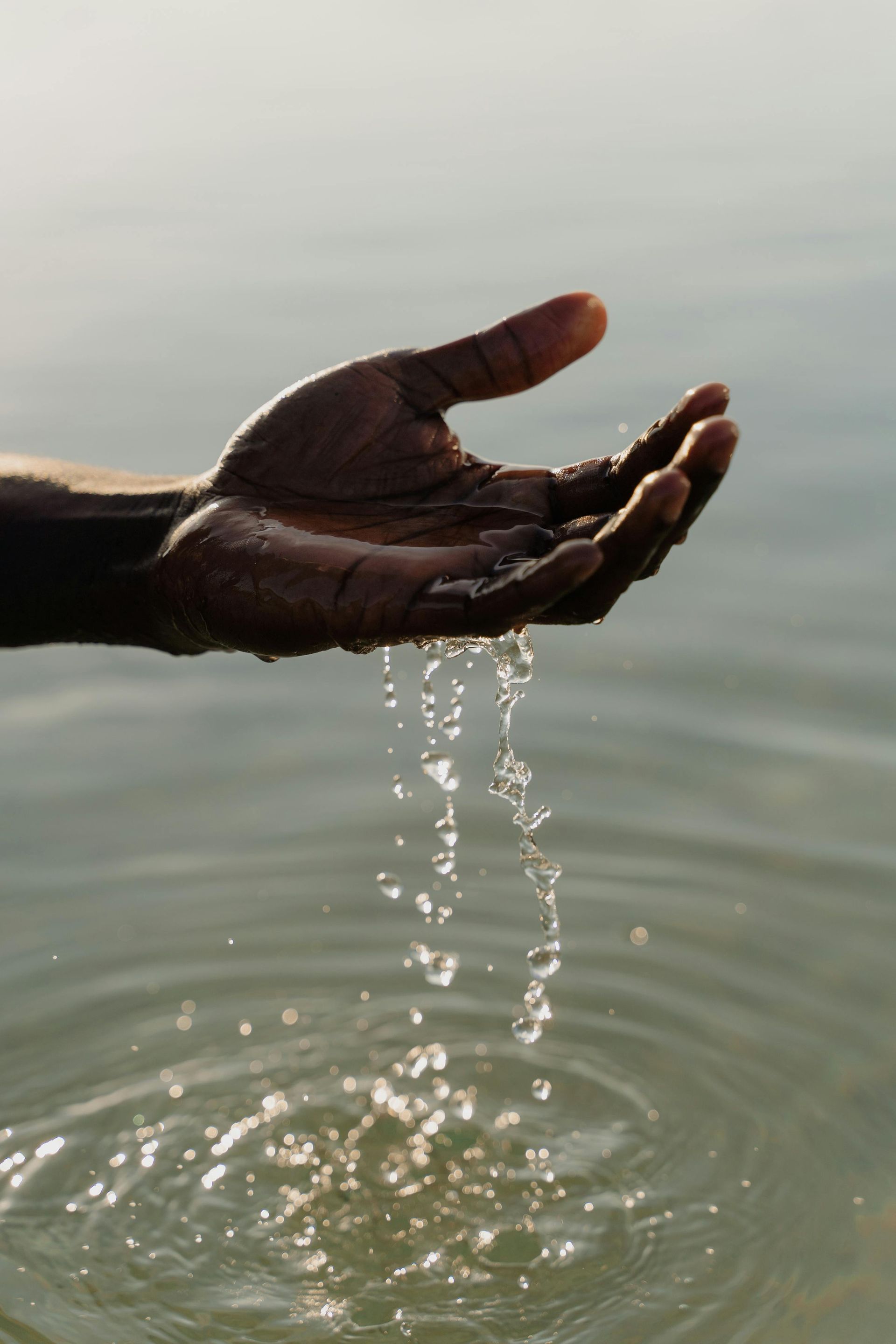 Black hand cupping water above a calm surface, with droplets streaming gently between fingers.